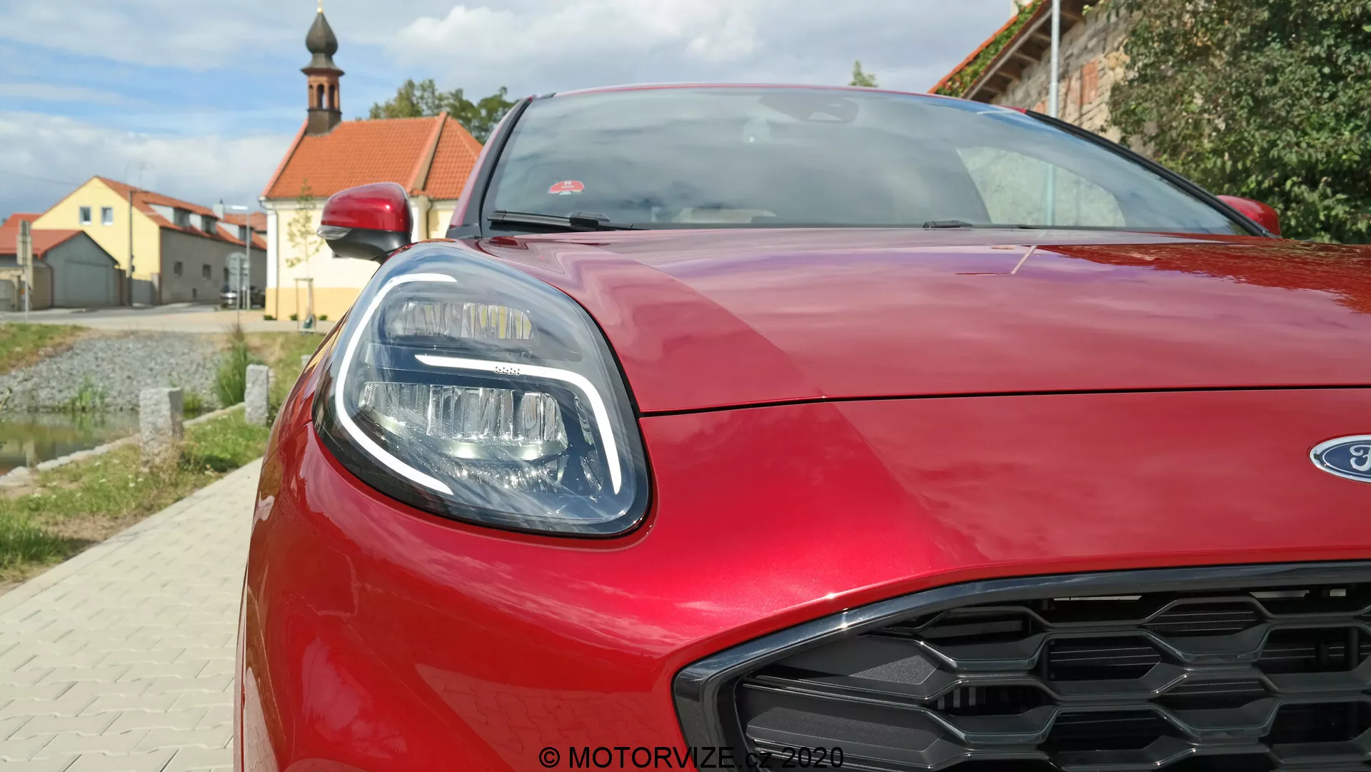 Detailný pohľad na prednú časť červeného auta Ford Puma 2020, ukazujúci pravý svetlomet s elegantnými LED prvkami, aerodynamicky tvarovanú kapotu a dizajn grilu v tvare včelieho plástu. Fotografia je urobená z nízkeho uhla a zachytáva moderný štýl a športové detaily vozidla.