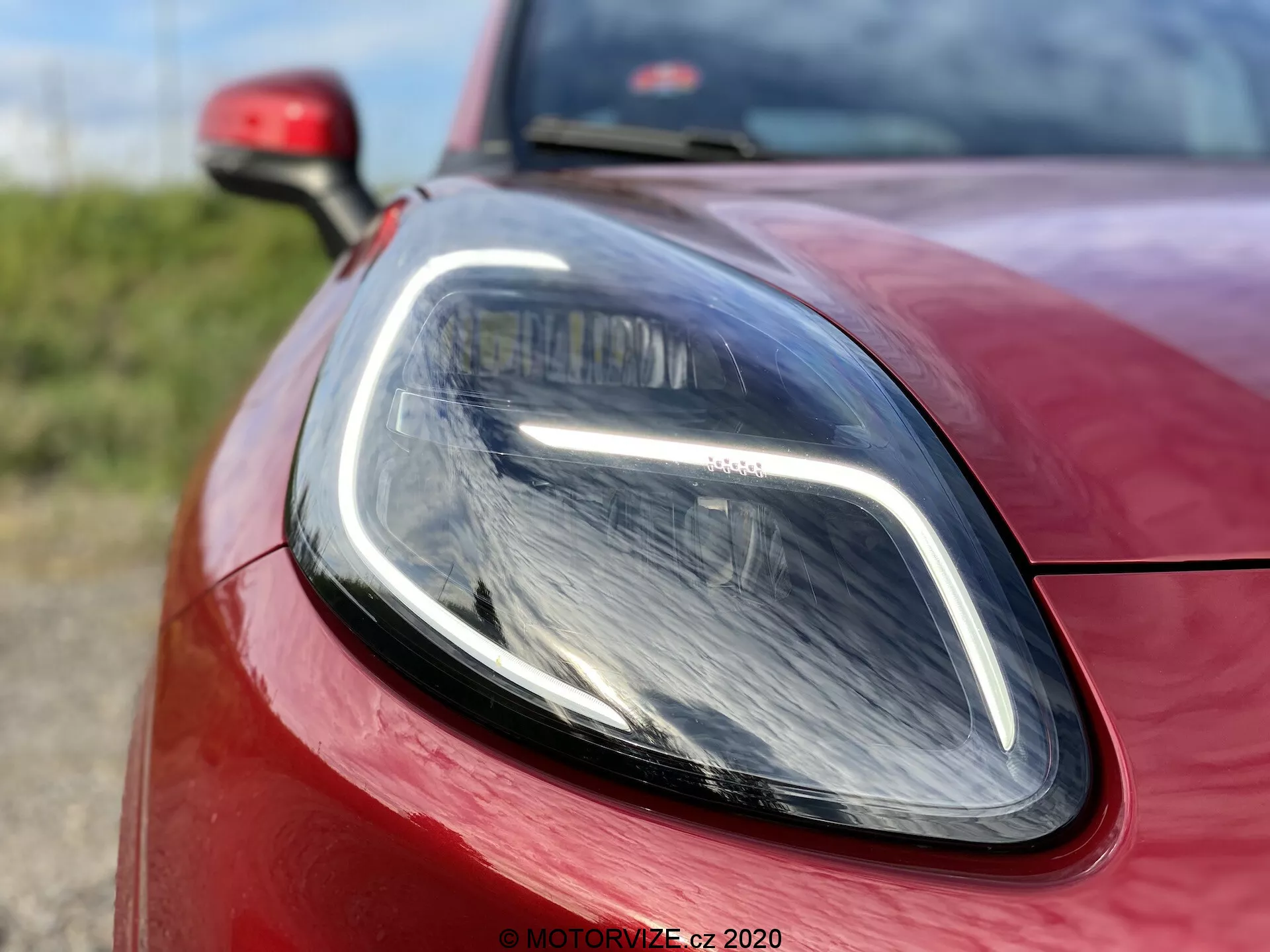 Detailný záber na Ford Puma 2020, zameraný na časť predného svetlometu na strane vodiča a okolitú karosériu. Fotografovaný z nízkeho, mierne posunutého uhla, zdôrazňuje elegantné denné LED svetlá a moderný projektorový svetlomet. Živá červená farba a presné zarovnanie panelov podčiarkujú športový, aerodynamický dizajn vozidla.