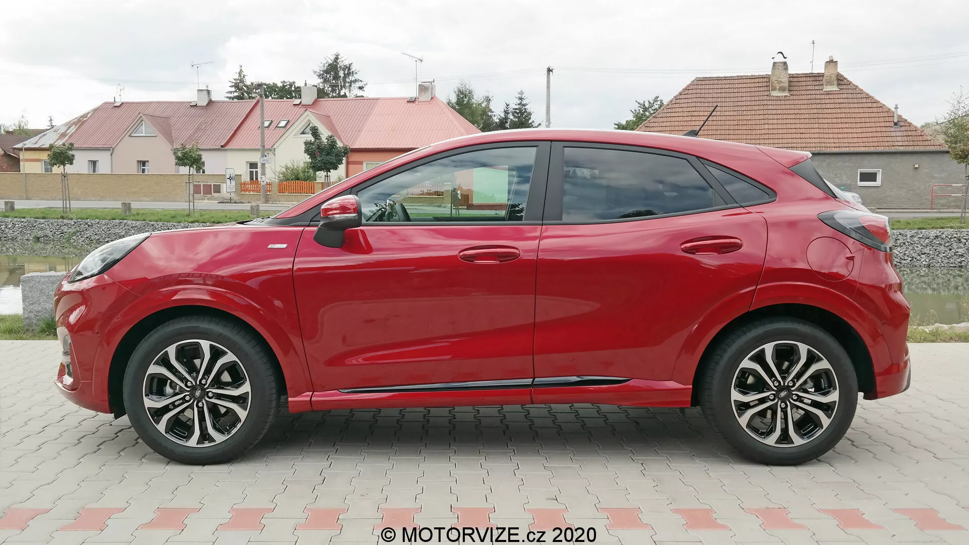 Bočný profil červeného, štvordverového hatchbacku Ford Puma 2020, nasnímaný počas dňa na parkovisku s trávou a obytnými budovami v pozadí. Auto má aerodynamické línie karosérie, viacramenné disky kolies, farebne zladené spätné zrkadlá a kľučky dverí a znižujúcu sa líniu strechy. Absencia viditeľných zámkov naznačuje možný bezkľúčový vstupný systém.