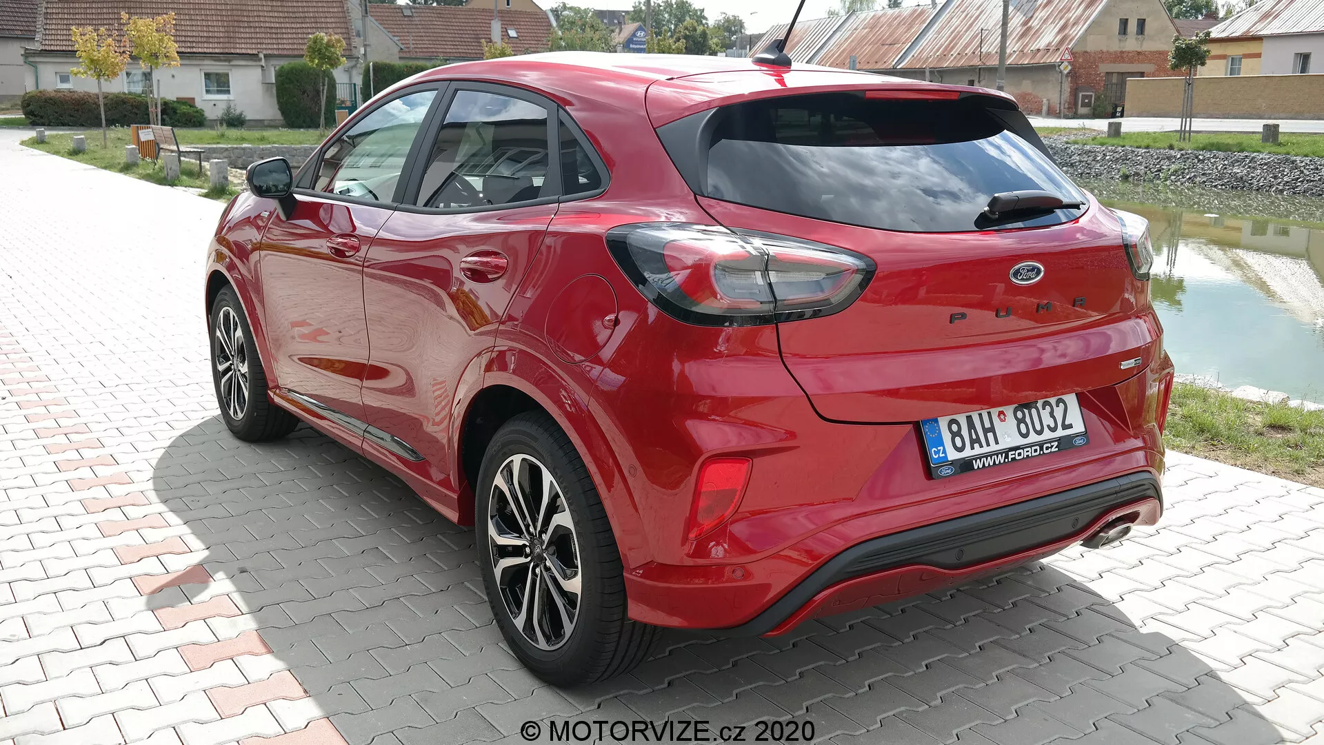 Pohľad na tri štvrtiny zadnej časti moderného hatchbacku Ford Puma 2020 v žiarivo červenej farbe, urobený za denného svetla. Fotografia zdôrazňuje aerodynamické kontúry vozidla, charakteristické zadné svetlá, ktoré obchádzajú boky, a integrované senzory zadného nárazníka, čo naznačuje možnosti parkovacieho asistenta. Dvojité výfukové hroty a zadný spojler zvyšujú športový vzhľad, zatiaľ čo viaclúčové zliatinové kolesá a prvky vo farbe karosérie, ako bočné zrkadlá a kľučky dverí, prispievajú k modernému dizajnu. Zadný stierač je viditeľný, čo naznačuje praktickosť v nepriaznivých poveternostných podmienkach.