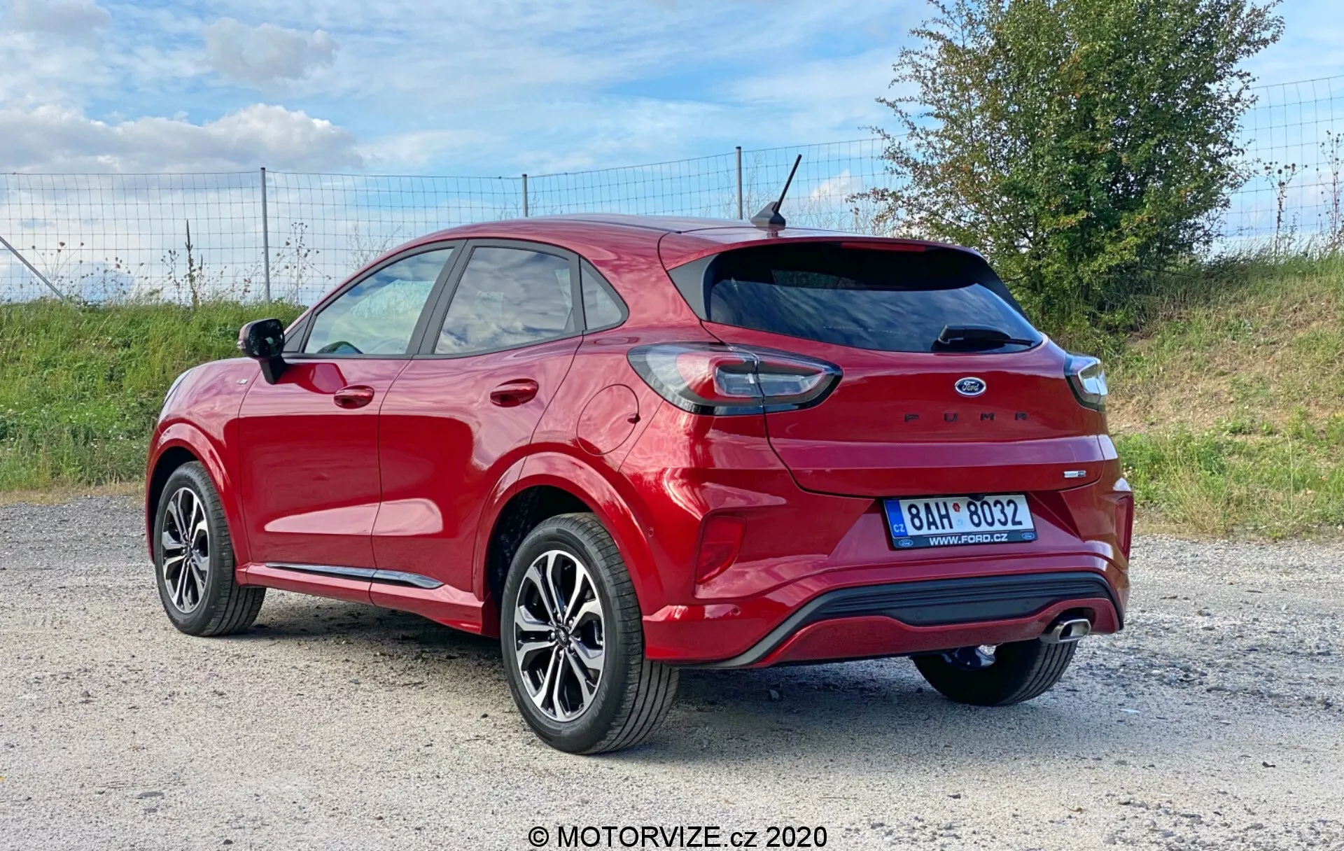 Obrázok predstavuje červený Ford Puma 2020 crossover/SUV vyfotografovaný zo zadného trojštvrťového uhla, ukazujúc zadné koleso a pneumatiku, časť predných dverí a zadných dverí pre pasažierov, zadný štvrťpanel a celý výklopný zadný diel s integrovaným spojlerom a zadným stieračom. Viditeľné prvky zahŕňajú zadné svetlá čiastočne obtočené okolo zadného blatníka, zadný nárazník s reflektormi a parkovacími senzormi, dvojitý výfukový systém, žraločiu anténu, zadné okno s odmrazovacími čiarami, liatinové kolesá s viacramenným dizajnom a pneumatiky s nízkym profilom, tónované okná, strešné lišty a púzdro spätného zrkadla ladiace s farbou karosérie. Vozidlo je situované v vonkajšom prostredí, možno na štrkovej alebo hlinenej ceste, s kúsok trávy v pozadí, čo naznačuje vidiecke alebo polovidiecke prostredie.