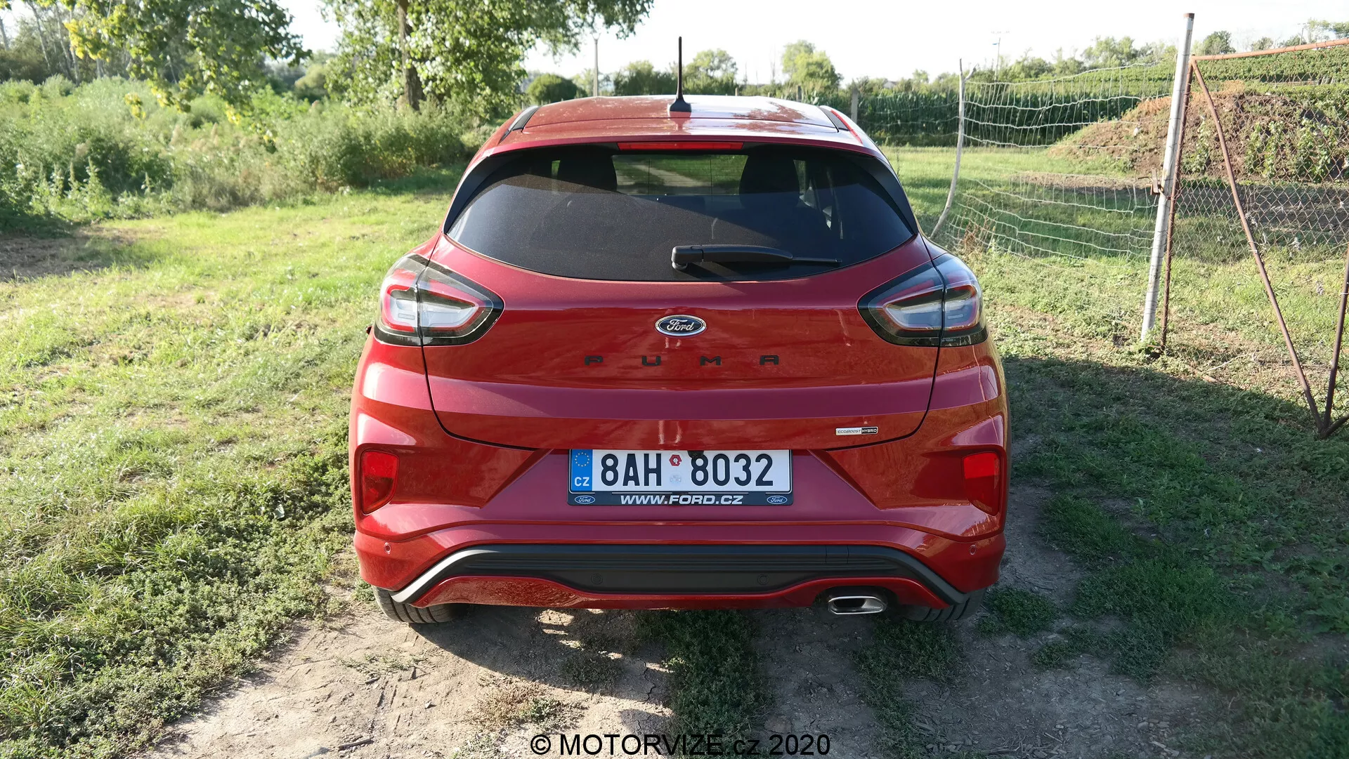 Pohľad na zadnú stranu červeného Forda Escape 2020 s hybridnou technológiou zaparkovaného na nespevnenom povrchu. Vozidlo má moderné zadné svetlá, centrálne umiestnené logo Ford a európsku registračnú značku. Zadné okno je vybavené stieračom a tretím LED brzdovým svetlom. Spodná časť karosérie a nárazník majú ochranné čierne plastové lišty s viditeľnými senzormi, ktoré naznačujú asistenta parkovania. Vegetácia v pozadí naznačuje vidiecke prostredie.