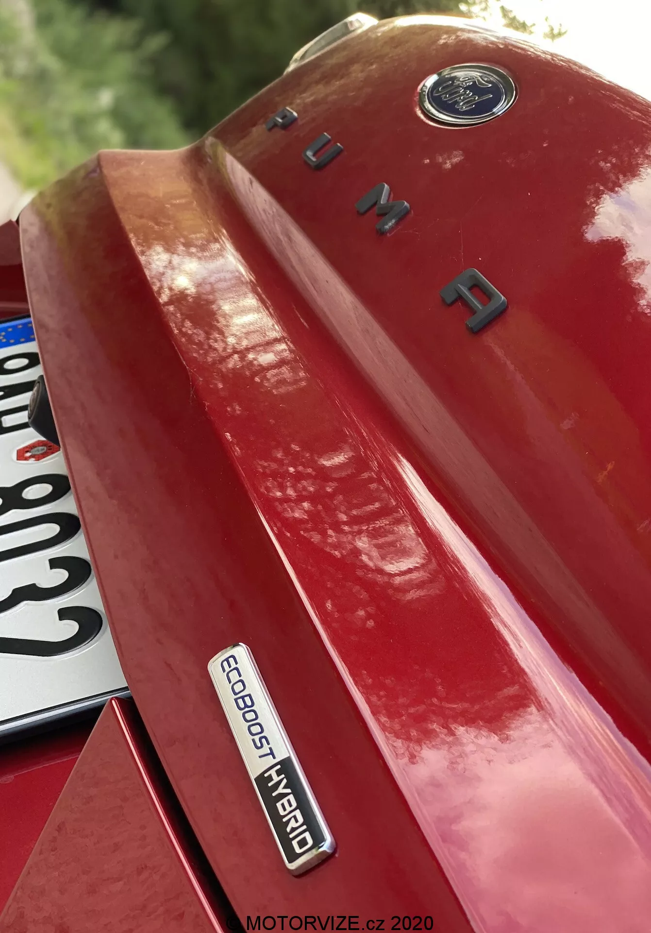 Detailný záber ľavej zadnej štvrtiny červeného vozidla Ford Puma 2020, odfotený zo stúpajúceho uhla, výrazne zobrazuje odznaky a značky. Obrázok zdôrazňuje emblém Ford na vrchu, názov modelu 'PUMA' v chrómových písmenách na kufri a odznak 'ECOBOOST HYBRID', čo naznačuje pokročilú hybridnú technológiu. Časť poznávacej značky je čiastočne viditeľná v ľavom dolnom rohu. Lesklý lak odráža svetlo a zvýrazňuje dizajn a technologickú sofistikovanosť vozidla.