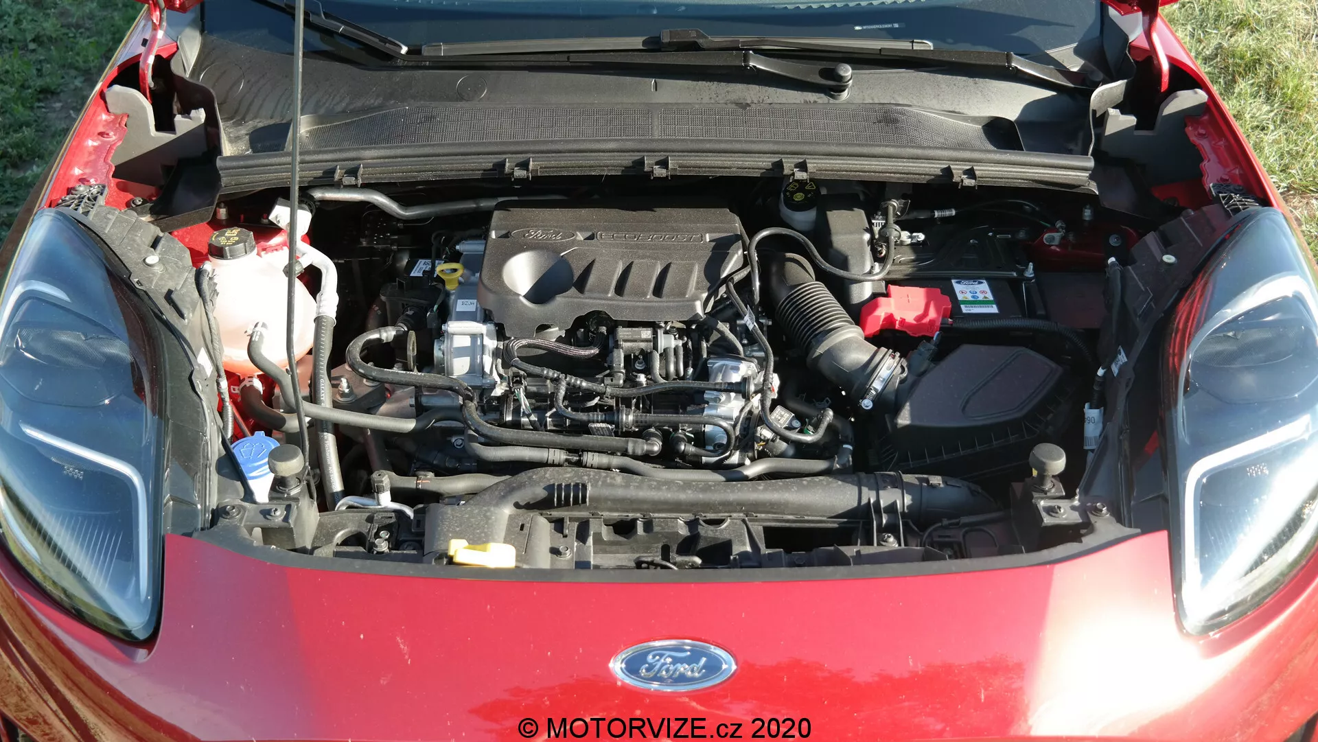 Detailný pohľad na motorový priestor Ford Puma 2020 s otvorenou kapotou, ukazujúci komponenty motora vrátane motorového bloku, krytu ventilov, sacieho potrubia a výfukového potrubia. Viditeľné sú aj prvky ako nádrže na kvapaliny, hadice, káble, batéria, vzduchové kanály a časť otvorenej kapoty s izoláciou.