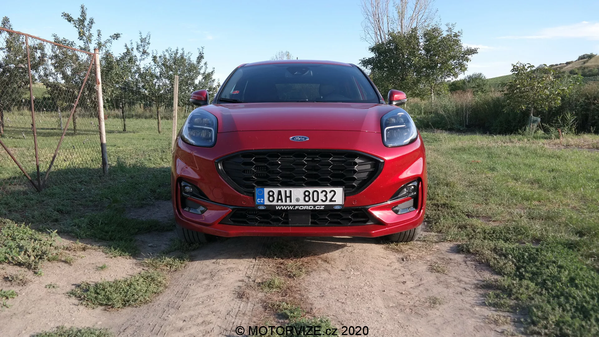 Predný pohľad na Ford Puma 2020, ktorý ukazuje jeho výraznú mriežku, elegantné LED svetlomety a aerodynamické línie kapoty. Auto je umiestnené na suchej pôde s trávnatými plochami, čo naznačuje vidiecke prostredie.