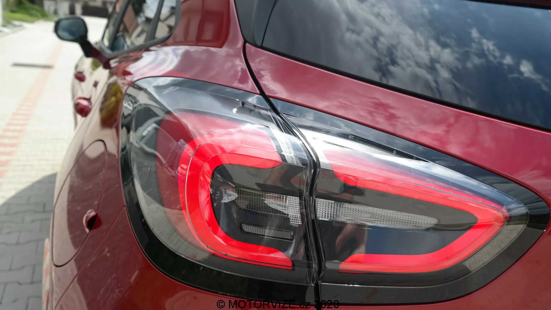 Detail zadnej časti vozidla Ford Puma 2020, zameraný na moderný dizajn LED zadných svetiel z mierne vyvýšeného uhla. Červené brzdové svetlo, číre a tmavšie segmenty pre spätné svetlo a smerové svetlá sa plynulo miešajú s hlbokým červeným alebo vínovým lesklým lakom vozidla, odrážajúc okolie. Obrázok zdôrazňuje zakrivený a moderný štýl vozidla a jeho funkčnú osvetľovaciu technológiu.