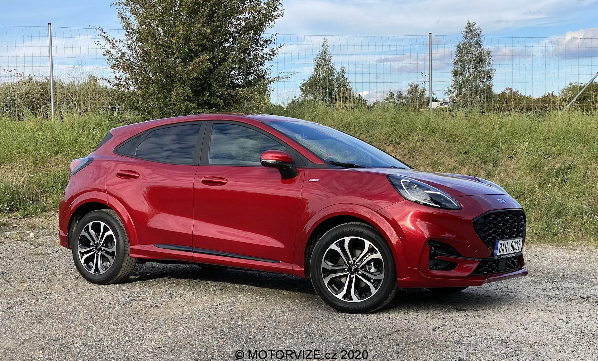 Profesionálna fotografia červeného kompaktného crossoveru SUV Ford Puma 2020, umiestneného na trávnatej ploche s lístím v pozadí. Bočný predný uhol zvýrazňuje výraznú prednú mriežku s dizajnom pripomínajúcim plást medu, moderné LED svetlomety a čiastočne viditeľné spätné zrkadlo. Športová, klesajúca línia strechy, výrazné línie dverí, jasne viditeľné kľučky, zliatinové kolesá s viacerými lúčmi a nízkoprofilové pneumatiky prispievajú k modernej estetike vozidla. Snímka, urobená na úrovni vozidla, zachytáva celú jeho dĺžku a zdôrazňuje moderný dizajn.