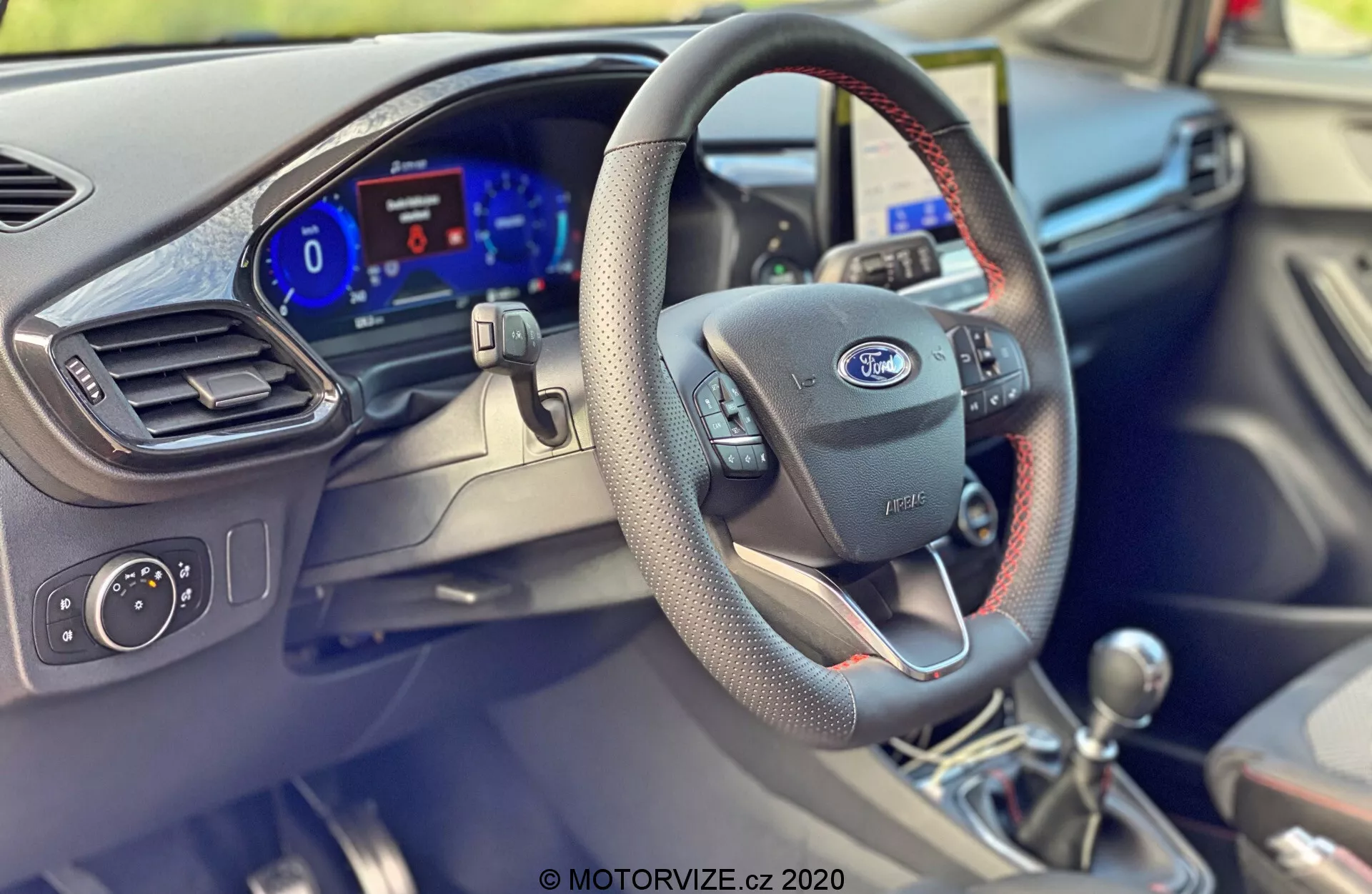 Interiér modelu Ford Puma 2020 z perspektívy úrovne očí zo sedadla spolujazdca, zdôrazňujúci stranu vodiča. Výrazné prvky zahŕňajú čierny volant s červeným prešívaním, nesúci logo Ford a moderné ovládacie tlačidlá. Na palubnej doske je viditeľný digitálny prístrojový panel s jasným modrým osvetlením. Gombík radiacej páky v pravom dolnom rohu, s prešívaním, sedí na radiacej páke manuálnej prevodovky. Časť dverí vodiča ukazuje ovládacie prvky na dverách, vzduchový ventil a ďalšie nastavenia, čo naznačuje sofistikovaný automobilový design a technológie.