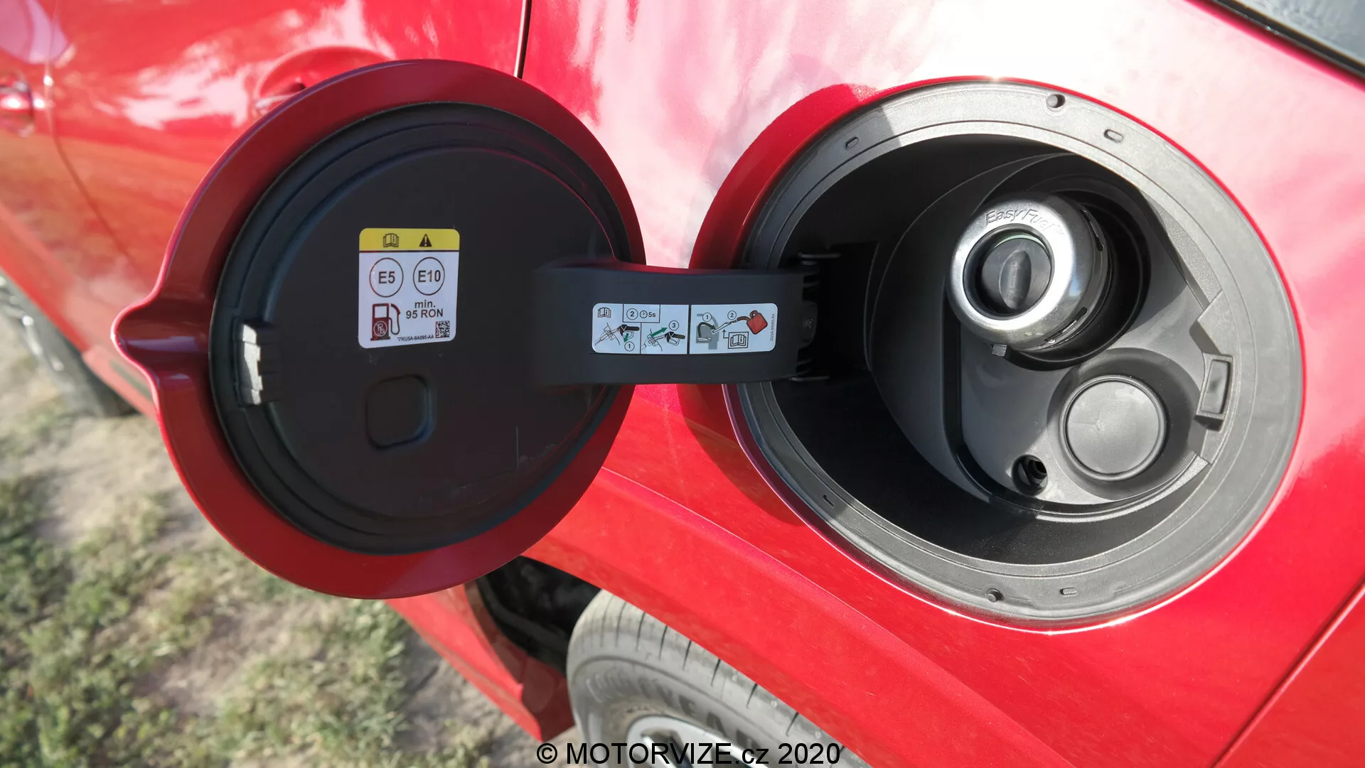 Detailný pohľad na otvorený nabíjací port typu 2 na žiarivo červenom elektrickom alebo plug-in hybridnom vozidle, zameraný na lesklú oblasť portu, závesnú krytku a informačnú nálepku viditeľnú vľavo vo vnútri dvierok portu, čo naznačuje dobre udržiavané auto s pokročilou technológiou elektrických vozidiel vonku.