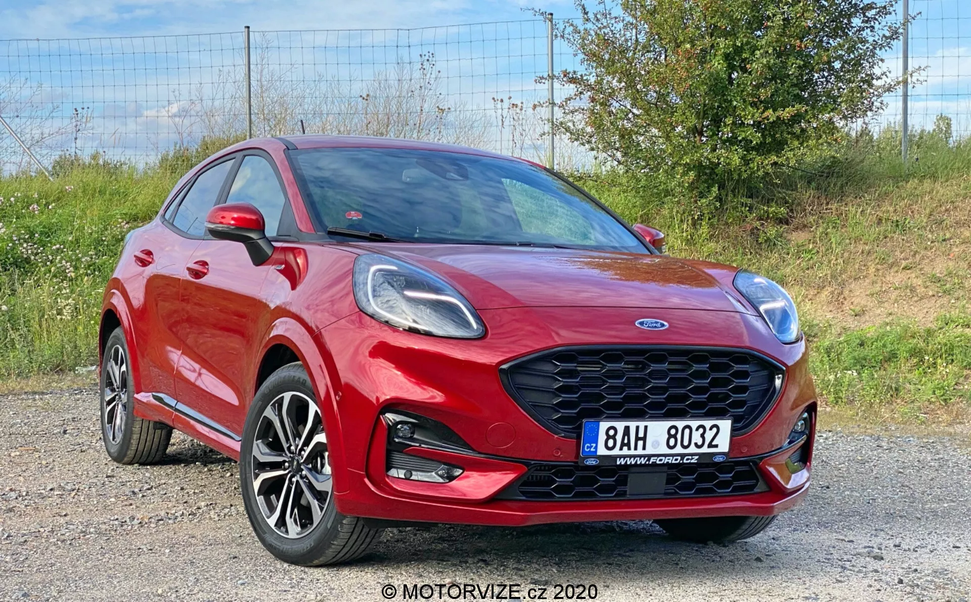 Obrázok zobrazuje žiarivo červený Ford Puma 2020, crossover zachytený z predného trojštvrtinového pohľadu, ktorý zobrazuje jeho elegantnú prednú mrežu s dizajnom včelieho plástu, uhlové svetlomety, viacramenné zliatinové kolesá, predný nárazník s integrovanými hmlovými svetlami a výraznú ramennú líniu. Dynamická kapota s záhybmi, zrkadlá s integrovanými smerovkami a časť predných a zadných dverí sú viditeľné, čo naznačuje konfiguráciu so štyrmi alebo piatimi dverami na pozadí štrkovej plochy s vegetáciou.