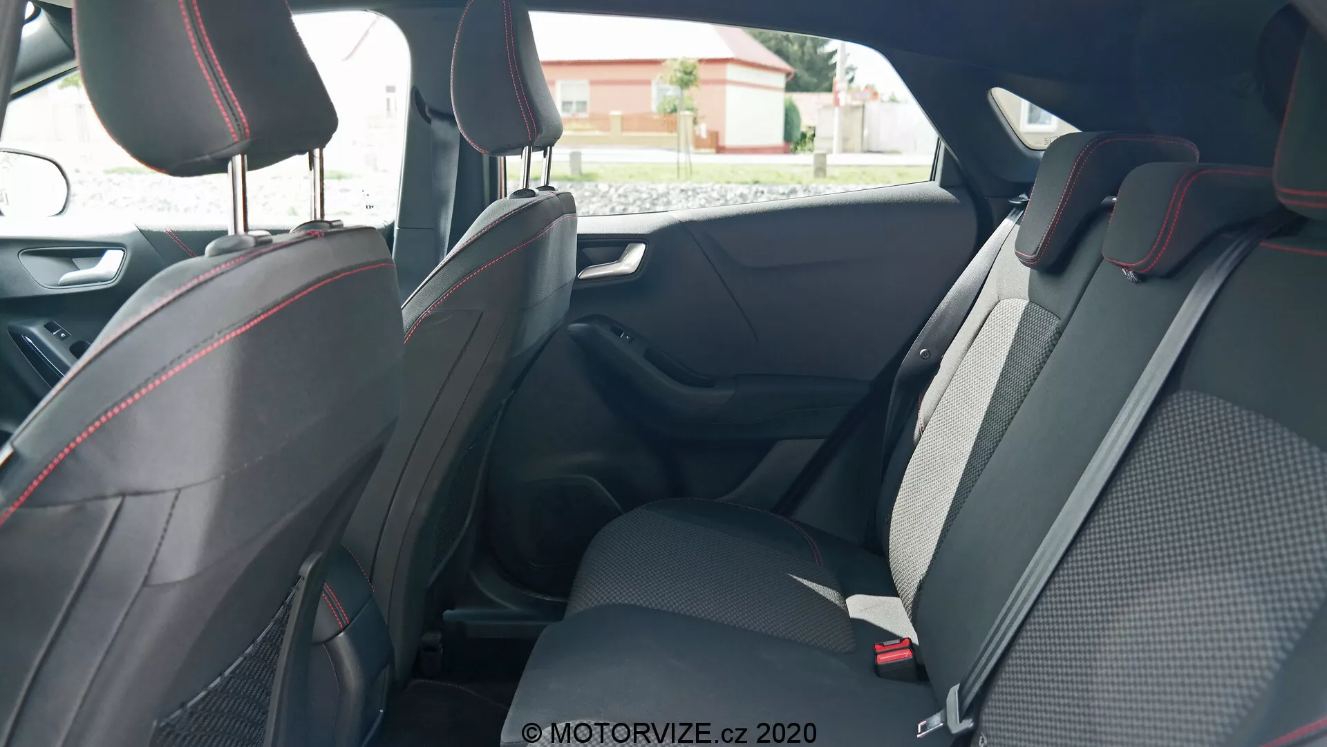 Interiér vozu Ford Puma 2020 zameraný na zadné sedadlá, vyhotovený zvonku zadných dverí pre cestujúcich, ukazujúci priestranný, športový dizajn s materiálmi z tkaniny a kože, zdôraznený červeným šitím. Viditeľné funkcie zahŕňajú nastaviteľné hlavové opierky, trojbodové bezpečnostné pásy a skladacie sedadlá pre lepšiu všestrannosť. Viditeľné sú aj dverové panely s rukoväťami, ovládaním elektrických okien a odkladacími priestormi, zdôrazňujúc ergonomické a pohodlné aspekty.