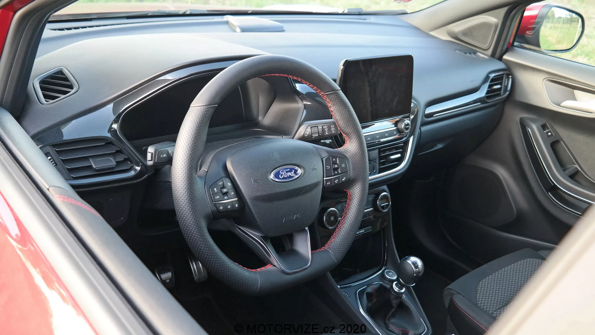 Pohľad na interiér kokpitu Ford Puma 2020 z mierne zvýšeného uhla, zameraný na stred palubnej dosky. Z pohľadu od dverí vodiča fotografia ukazuje trojramenný volant s multifunkčnými tlačidlami pre ovládanie audia, hlasových príkazov a tempomatu. Uprostred volantu je výrazne viditeľné logo Ford. Za volantom je viditeľná časť prístrojového panela. Stred palubnej dosky obsahuje infotainment systém, pravdepodobne s navigáciou, prehrávaním médií a nastaveniami. Nižšie je manuálne ovládanie klímy s otočnými ovládačmi a tlačidlami pre teplotu, klimatizáciu a odmrazovanie. Na obrázku sú viditeľné aj voliaci páka automatickej prevodovky a ručná brzda. Interiér kombinuje tmavé plasty na vrchnej časti palubnej dosky s čiernou látkou alebo kožou na spodných častiach, čo prezentuje funkčný dizajn zameraný na používateľnosť.