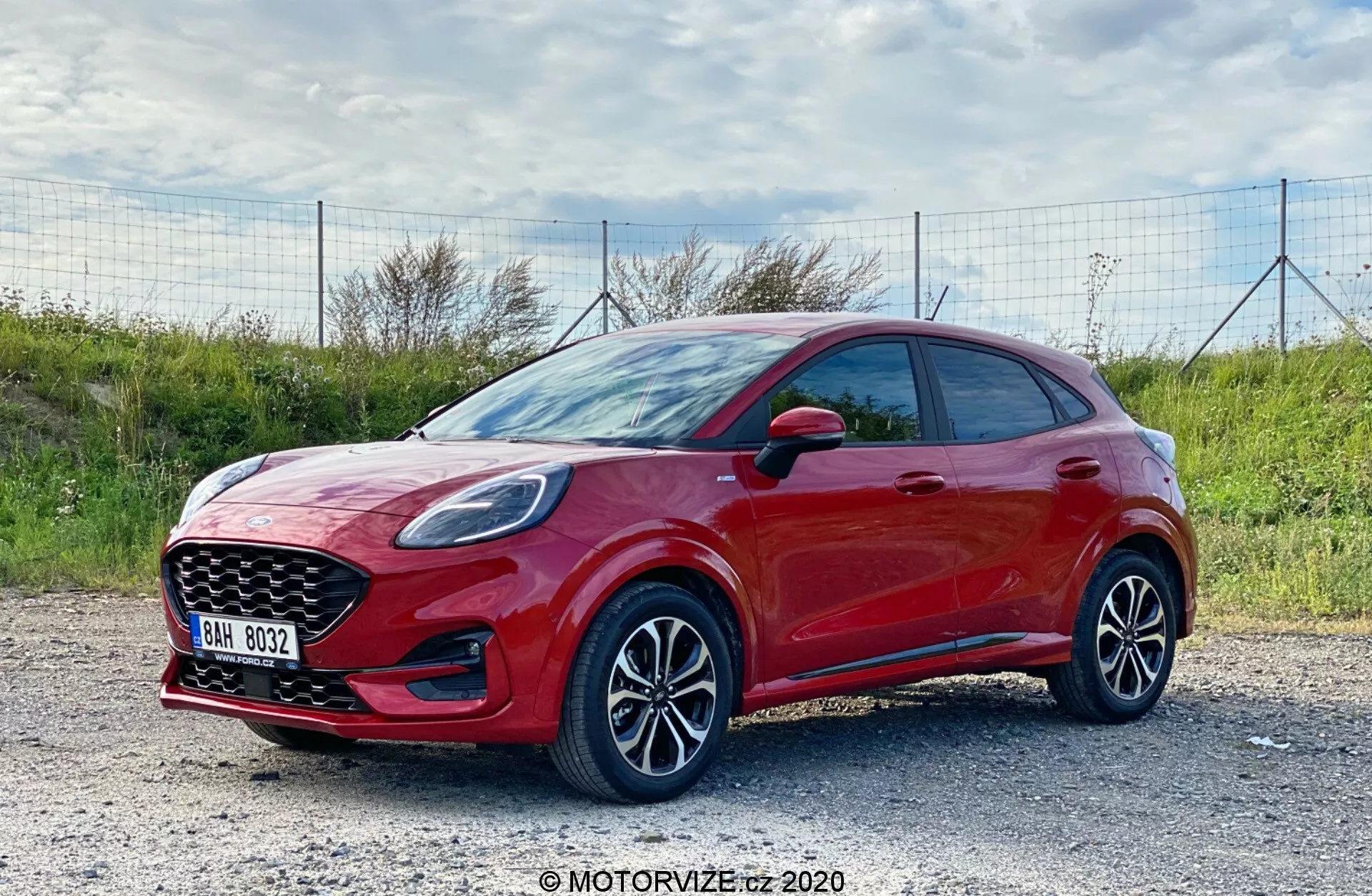 Bočný profil moderného hatchbacku Ford Puma 2020, zachytený za denného svetla s prírodným vonkajším pozadím. Vozidlo v žiarivej červenej farbe s lesklým povrchom je viditeľné z uhla tri štvrte zpredu, jasne ukazujúc svoj bočný profil a prednú časť. Auto je zaparkované na štrkovom povrchu. Záber, urobený mierne nad úrovňou kolesa, zdôrazňuje viaclúčové hliníkové disky a spodnú časť vozidla. Viditeľné sú elegantné predné svetlá pravdepodobne s LED technológiou, výrazná predná mriežka a bočné zrkadlá s integrovanými indikátormi. Auto má kľučky dverí vo farbe karosérie, bez viditeľných ochranných bočných líšt a športový štýl hatchbacku so znižujúcou sa líniou strechy. Technologické funkcie pravdepodobne zahŕňajú pokročilé asistenčné systémy pre vodiča a infotainment s možnosťami pripojenia.