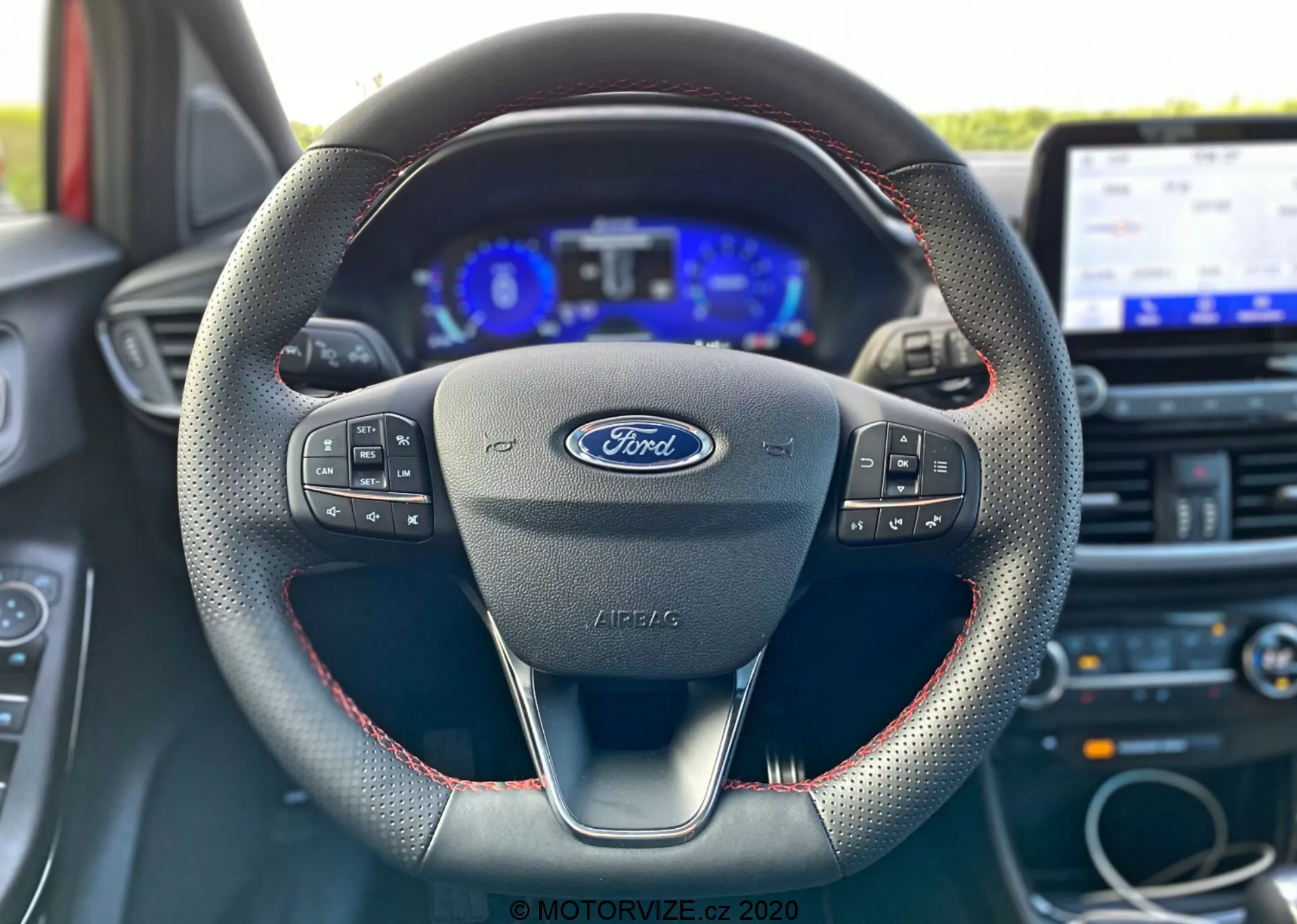 Interiérový záber Fordu Puma 2020 z pohľadu vodiča, zameriavajúci sa na volant s logom Ford a multifunkčnými ovládačmi, moderným dizajnom s tromi ramenami s červeným prešívaním naznačujúcim kožený materiál, digitálnou palubnou doskou lemovanou analógovými ukazovateľmi a dotykovým infotainment systémom v pozadí.