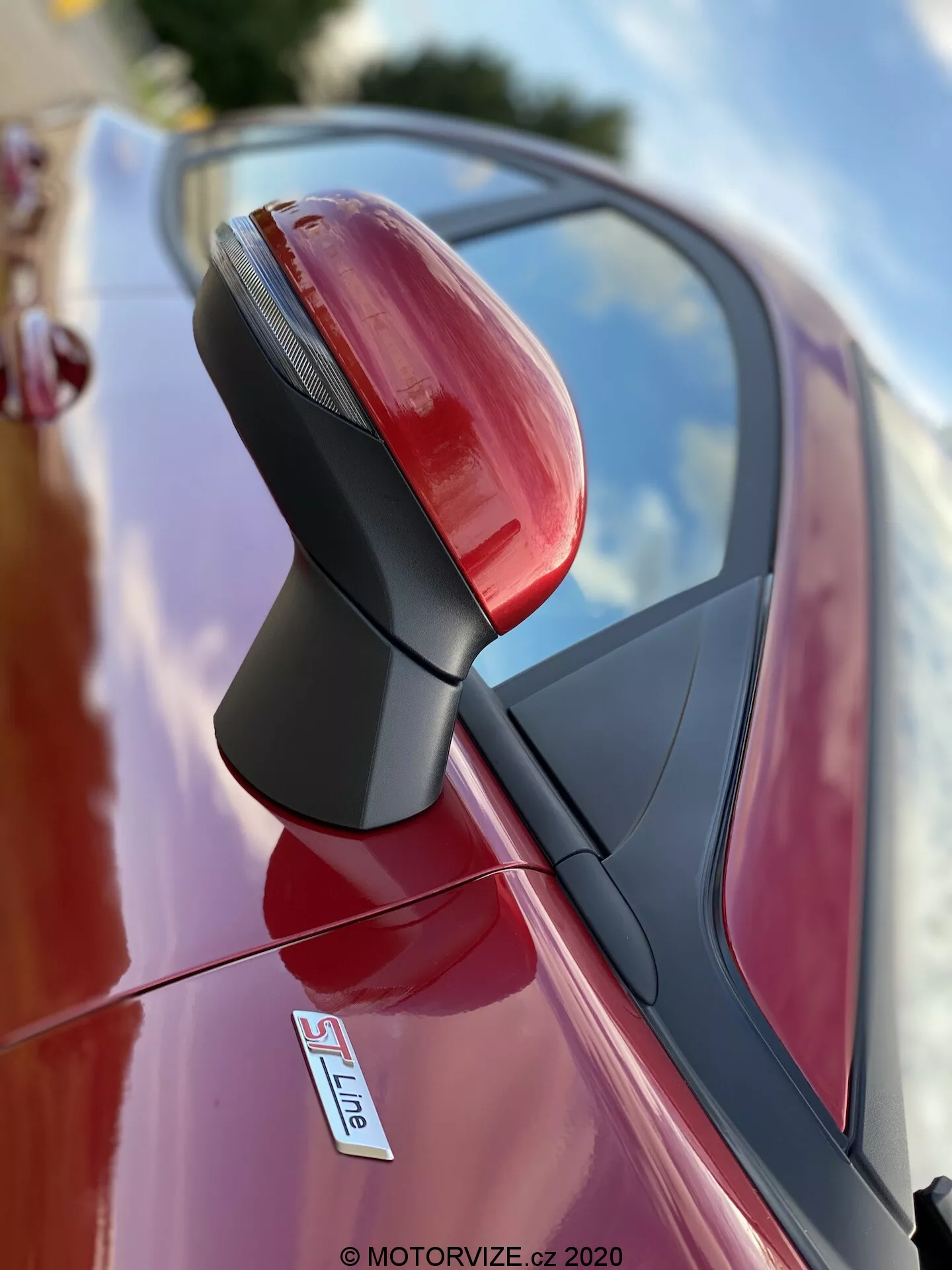 Detailný záber na vonkajšie spätné zrkadlo na strane vodiča červeného auta, pravdepodobne Audi so znakom S Line. Fotografia vyhotovená z nízkeho uhla sa zameriava na zostavu zrkadla s malou hĺbkou ostrosti, čím rozmazáva pozadie. Zrkadlo je ostré a detailné, zobrazuje integrované smerovky, textúrovanú čiernu podporu a lesklý, kovový povrch. Odrazy oblohy a okolia naznačujú, že auto je zaparkované vonku.