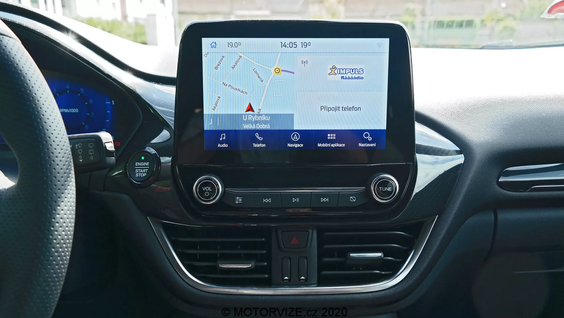 Stredová konzola interiéru Ford Puma 2020 z pohľadu vodiča alebo spolujazdca, zobrazujúca infotainment systém s dotykovou obrazovkou, ktorá ukazuje navigačnú mapu, fyzické tlačidlá pre varovné svetlá, nastavenie klimatizácie, CD slot, USB port a 12V napájaciu zásuvku. Konzola tiež zahŕňa digitálny teplomer, ktorý ukazuje 17°C, a hodiny, ktoré ukazujú 14:05, všetko obklopené modernými prvkami interiéru so zakrivenými líniami a ventilačnými otvormi klimatizácie.