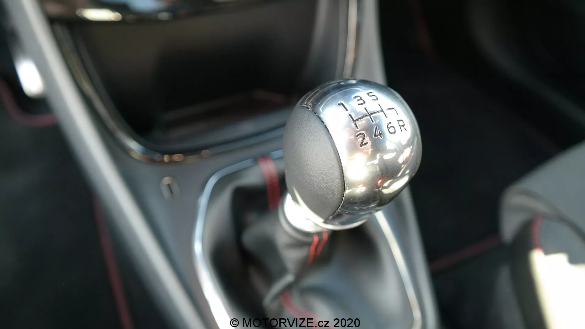 Detailná fotografia manuálnej radiacej páky vozidla Ford Puma 2020, fotografovaná z mierne zvýšeného uhla, ktorý ukazuje horný a bočný profil. Kovová hlavica radiacej páky má čierny vrch s bielymi vyrytými číslicami, ktoré indikujú vzor 6-stupňovej prevodovky s písmenom „R“ pre spiatočku. Radiaca páka je namontovaná na base s chrómovým povrchom a pokrytá čiernou manžetou, pričom ostré zameranie fotoaparátu je na hlavici radiacej páky, zatiaľ čo pozadie je rozmazané, čo zvýrazňuje modernú a štýlovú estetiku interiéru vozidla a technológiu pohonnej jednotky.