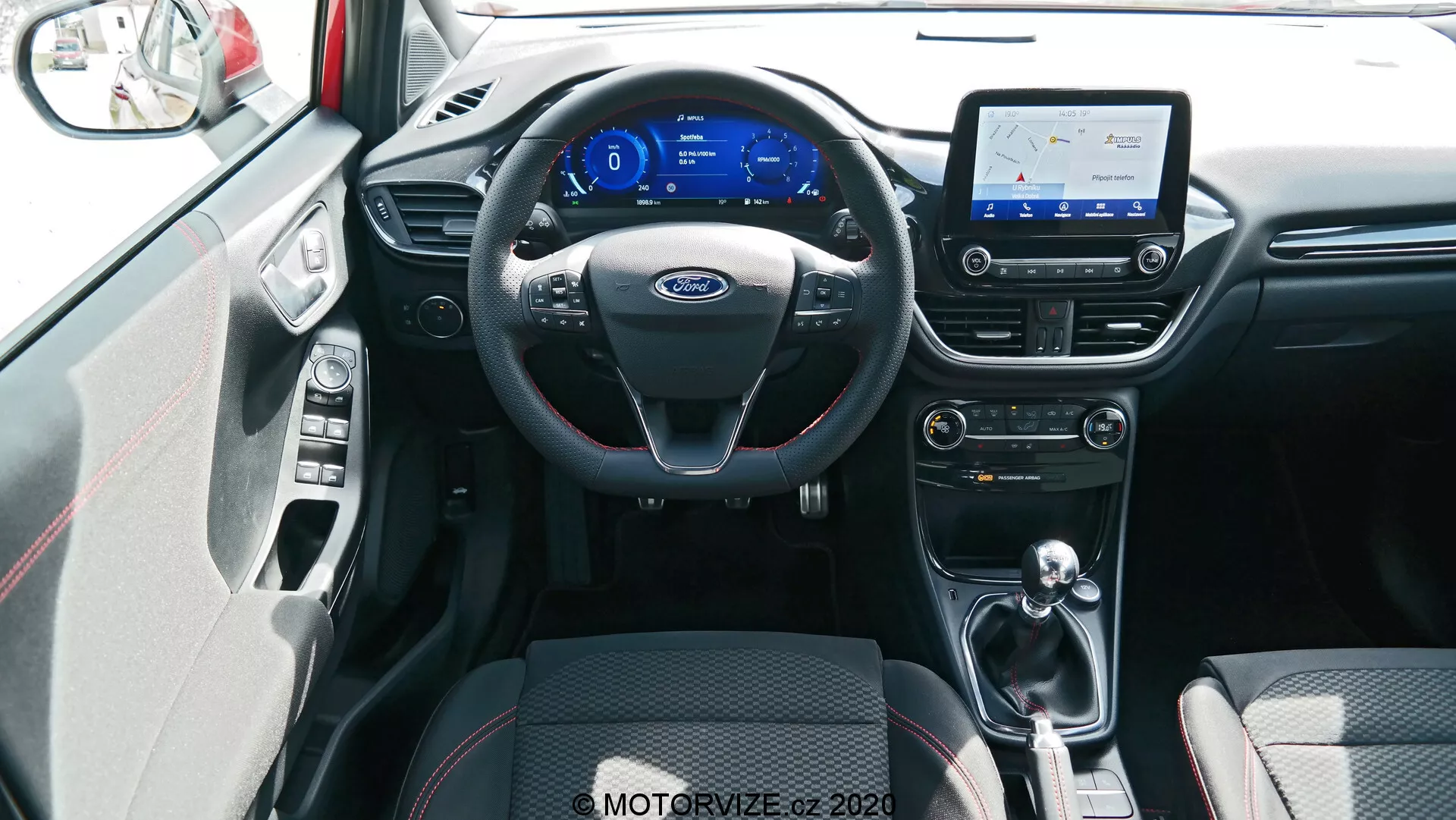 Pohľad do interiéru vozidla Ford Puma 2020, zhotovený z centrálnej pozície zadného sedadla. Obrázok zobrazuje čierny trojramenný volant s multifunkčnými ovládačmi a striebornými akcentmi, digitálnu prístrojovú dosku s modrou témou a infotainmentový dotykový displej zobrazujúci mapu, čo naznačuje navigačné schopnosti. Pod displejom sú ovládacie prvky klimatizácie a ventilácie s otočnými voličmi a tlačidlami na nastavenie teploty a rýchlosti ventilátora. Stredový tunel obsahuje radiacu páku, čo naznačuje automatickú alebo automatizovanú manuálnu prevodovku. Predné sedadlá sú potiahnuté čiernym látkovým čalúnením s dvojfarebným vzorom pre pohodlie a úchop, viditeľná je aj časť vnútorného panelu dverí so striebornou kľukou a zodpovedajúcimi akcentmi. Podlaha je pokrytá kobercom, ktorý ladí s farebným schémou interiéru, so zrejme dobre viditeľným prostredím osvetlenia. Záber je zhotovený z mierne vyvýšeného uhla, ktorý komplexne zobrazuje predný interiér a dostupné technologické prvky.