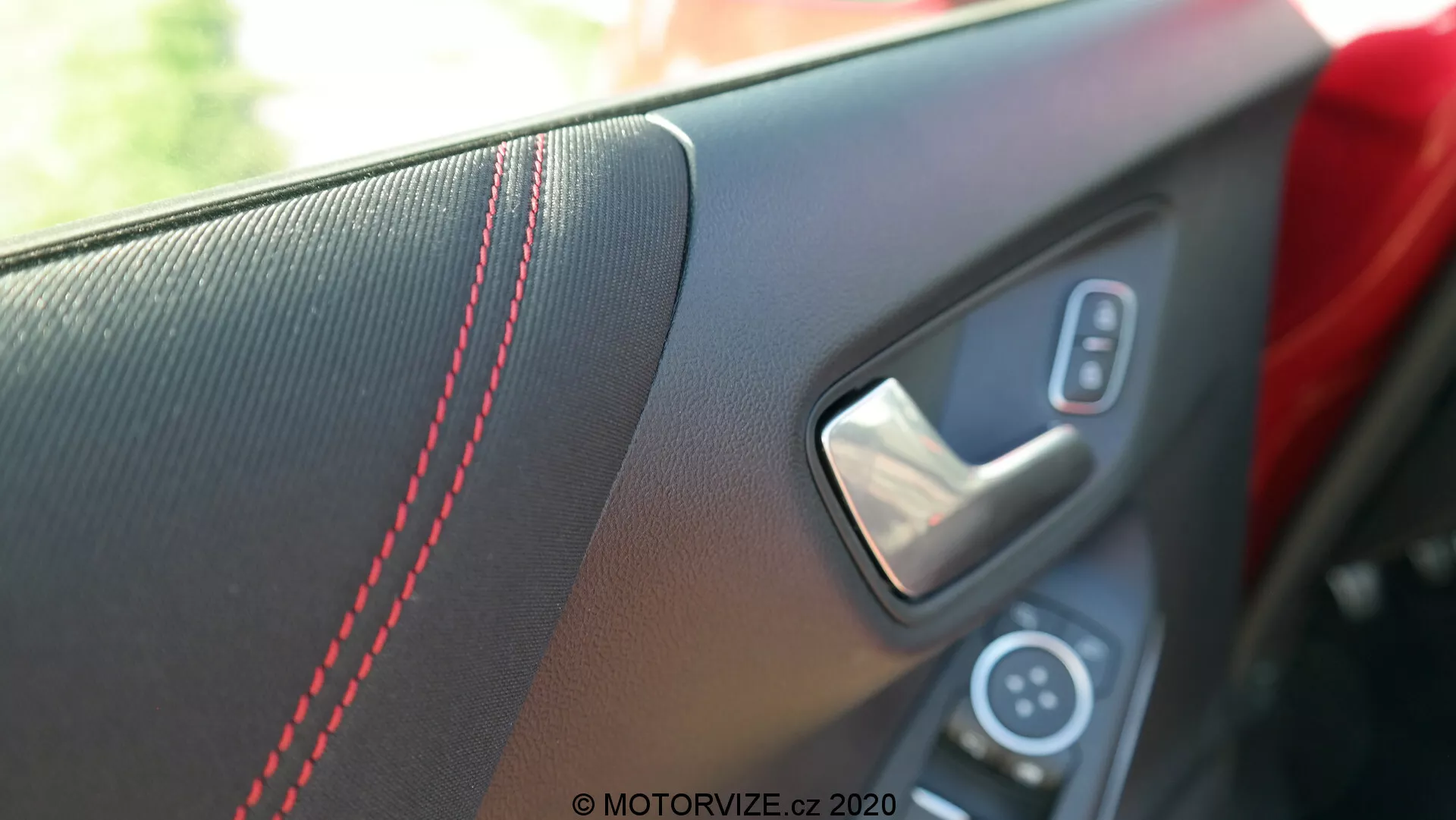 Detailný pohľad na vnútorný panel dverí Ford Puma 2020 z perspektívy cestujúceho alebo vodiča vo vnútri vozidla. Obrázok sa zameriava na oblasť okolo kľučky, zvýrazňujúc textúrovanú vložku s červeným kontrastným prešívaním, kovovú kľučku dverí a časť ovládacieho panelu s tlačidlom elektrického ovládania okien. Plytká hĺbka ostrosti rozmazáva pozadie a zdôrazňuje športové a luxusné dizajnové prvky v prirodzenom dennom svetle.