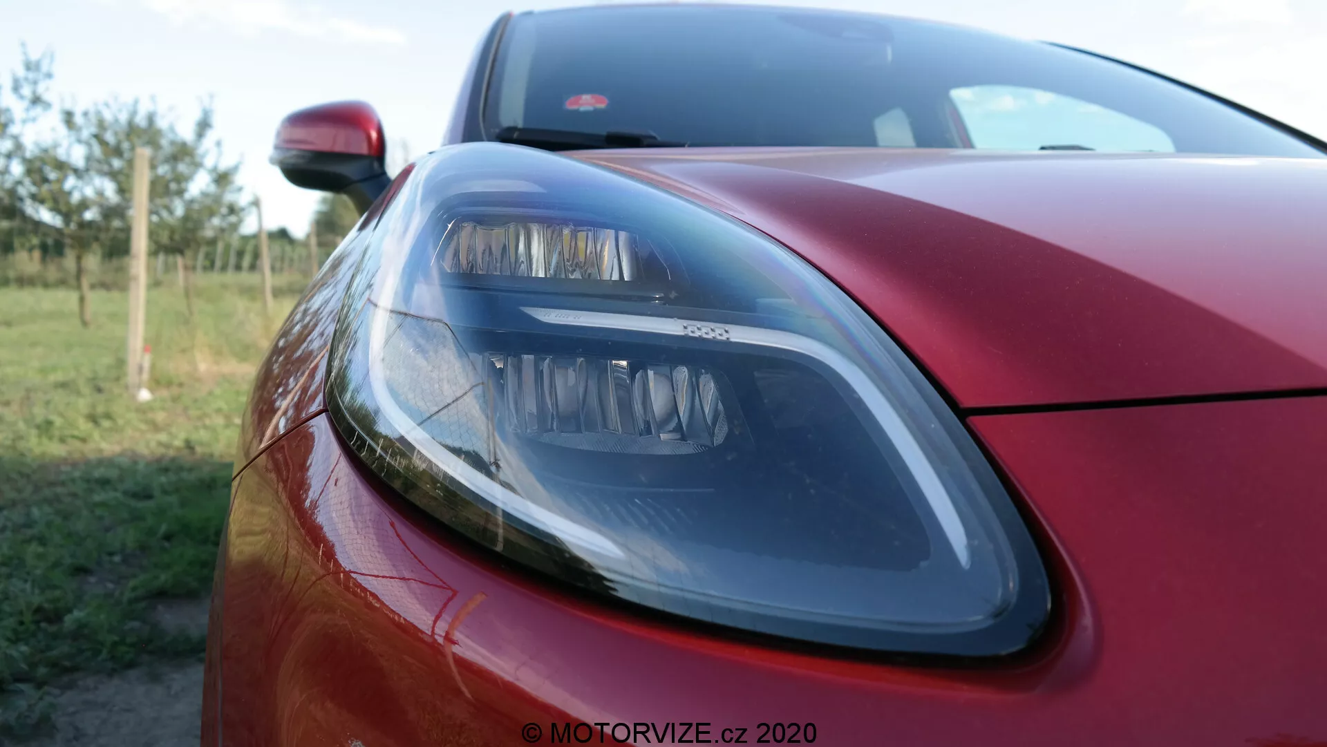 Detailný záber na prednú ľavú časť vozidla Ford Puma 2020 vo farebnom odtieni hlboká metalická červená, zobrazujúci moderný svetlomet s elegantnými dizajnovými prvkami, časť kapoty a pohľad na prednú mriežku s malou hĺbkou ostrosti rozostrujúcou pozadie, zvýrazňujúci prémiovú povrchovú úpravu a technológiu osvetlenia.