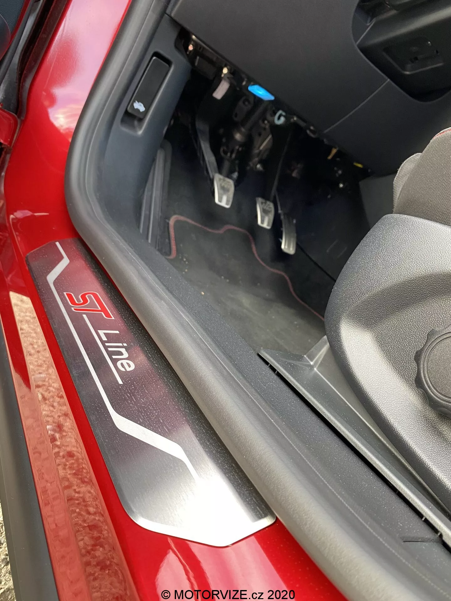 Detailný pohľad na prah dverí vozidla Audi s označením 'S Line' v striebornej a červenej farbe na čiernom pozadí. Záber zhora a mierne dovnútra zachytáva spodný rám dverí, vnútornú hranu a časť priestoru na nohy vodiča s viditeľnými pedálmi brzdy a plynu. Prirodzené denné svetlo osvetľuje scénu, zvýrazňujúc červenú farbu exteriéru a komponenty interiéru v blízkosti palubnej dosky.