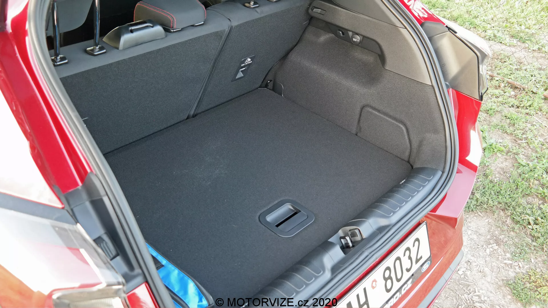Obrázok ukazuje zadný priestor alebo kufor automobilu Ford Puma 2020. Fotografia je urobená z uhla mierne zhora a zboku, čo poskytuje jasný pohľad na priestranný interiér kufra a jeho prah. Tmavý koberec naznačuje, že je navrhnutý tak, aby minimalizoval vzhľad nečistôt. Nie sú viditeľné žiadne zavazadlá, čo zdôrazňuje čistotu a priestrannosť kufra. Viditeľné prvky zahŕňajú bočné zárezy alebo vrecká na uloženie drobných predmetov a háčik alebo upevňovací bod na zabezpečenie predmetov. Čiastočne je vidieť zadný svetelný cluster auta na pravej strane obrázka.