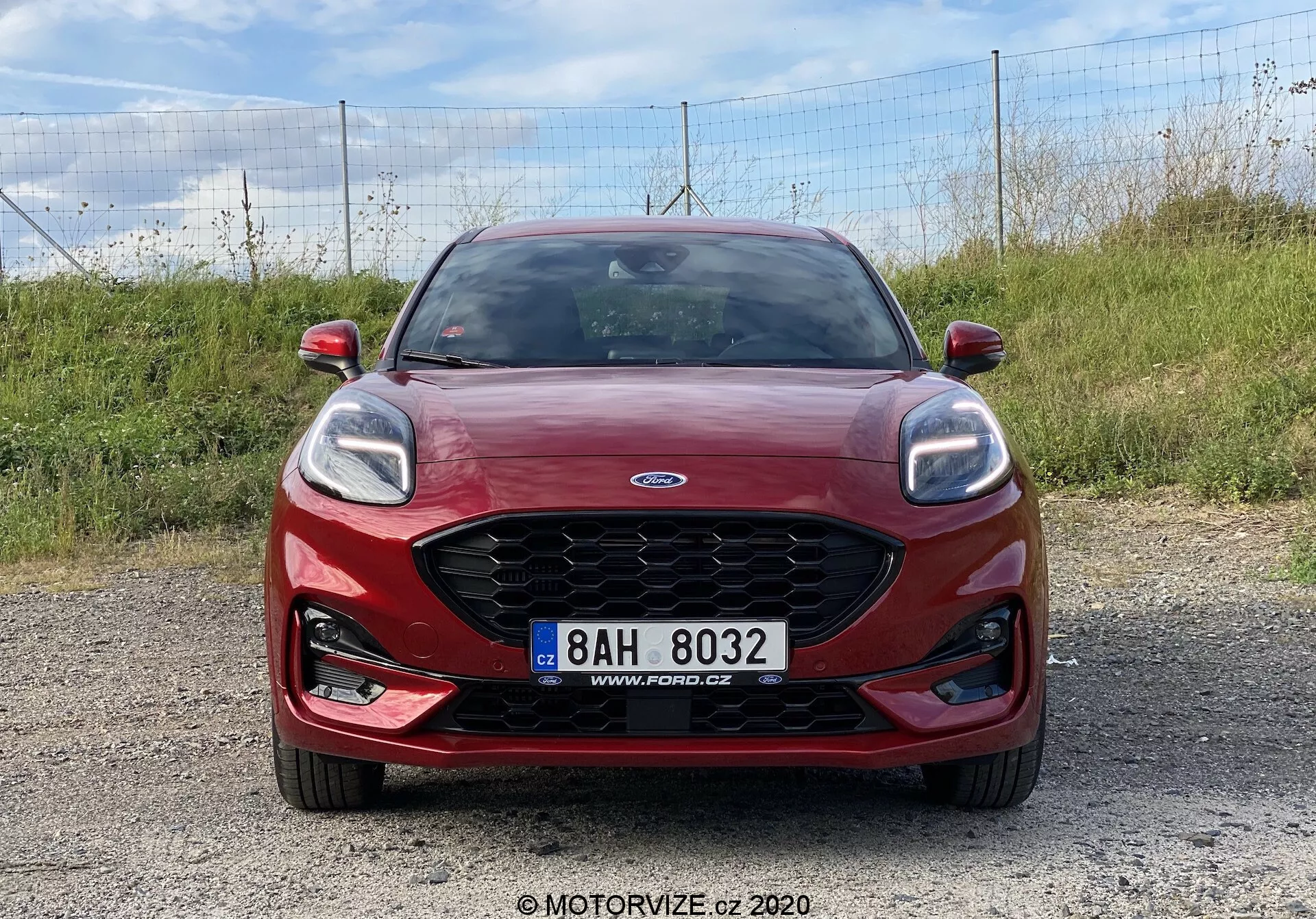 Červený Ford Puma 2020 zachytený z čelného pohľadu na štrkovom povrchu, ktorý ukazuje charakteristickú mriežku Ford s centrálnym logom, elegantné svetlomety s LED dennými svetlami, integrovanými hmlovými svetlami a centrálne umiestnenou registračnou značkou. Ideálne pre dizajn prednej časti vozidla, identifikáciu značky a moderné osvetľovacie systémy automobilov.