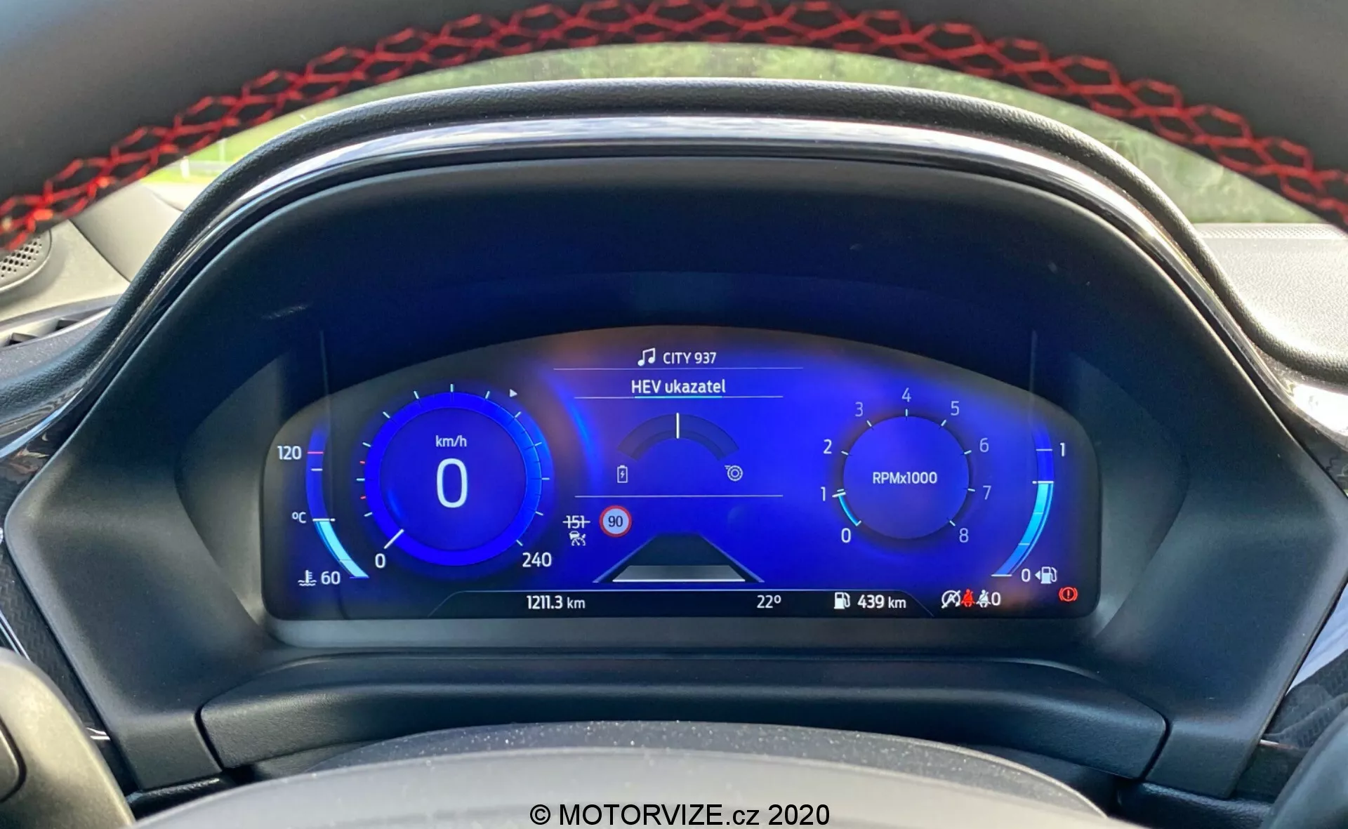 Detailný pohľad na moderný digitálny prístrojový panel Ford Puma 2020 z perspektívy vodiča. Digitálny displej obsahuje rýchlomer s ekologickými rýchlostnými rozsahmi vľavo, otáčkomer v strede, hybridné ukazovatele paliva vpravo a ďalšie informácie ako najazdené kilometre, vzdialenosť cesty, dojazd a vonkajšiu teplotu dole. Hore v strede sa zobrazuje oznámenie 'HEV úzkazník!', ktoré naznačuje režim hybridného elektromobilu. Elegantný, technologicky zameraný dizajn v odtieňoch modrej, bielej a červenej zdôrazňuje pokročilé informačné systémy pre vodiča a hybridnú technológiu vozidla.