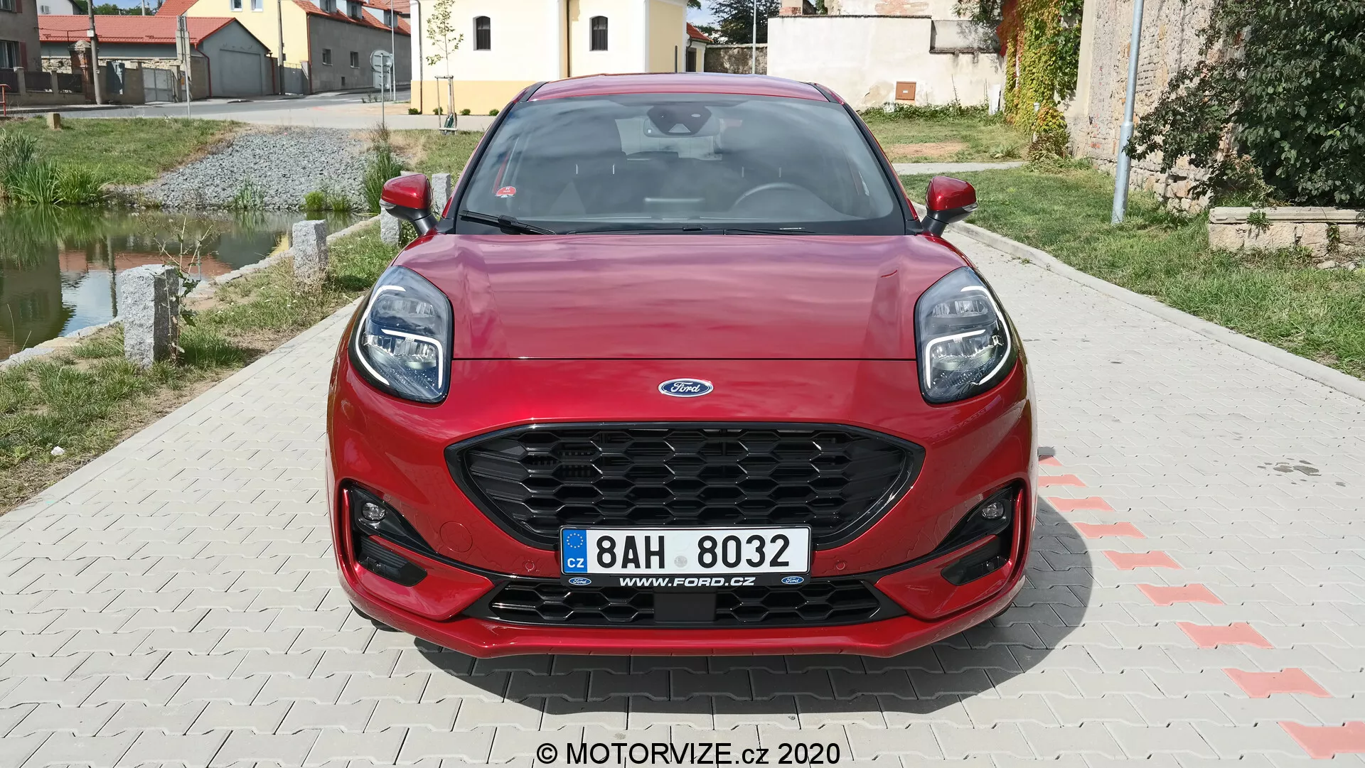 Predný uhol pohľadu na červený Ford Puma 2020, zobrazujúci šesťuholníkovú mriežku s čiernou sieťovinou, moderné prepadnuté svetlomety, športový predný nárazník so širokými nasávacími otvormi a centrálnu značku. Dynamické línie kapoty zvyšujú estetiku vozidla. V pozadí je vidieť obývateľnú oblasť.
