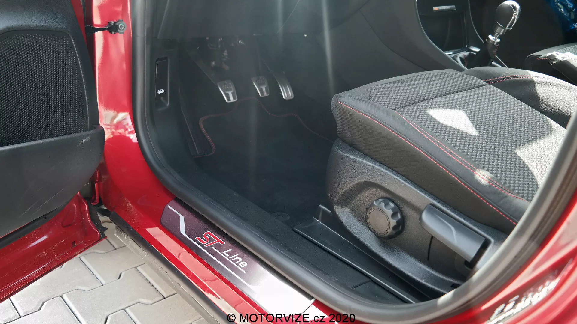 Detailný pohľad na interiér Fordu Puma 2020, sústrediaci sa na kovový prah dverí s označením S Line, športové sedadlo s kombináciou kože a textilu s bočnými opierkami, elektronické ovládanie sedadla, kovové pedály s gumenými vložkami pre manuálnu prevodovku, kobercovú rohož, červeno-čierny interiérový panel s mriežkou reproduktora a priame slnečné svetlo osvetľujúce interiér.