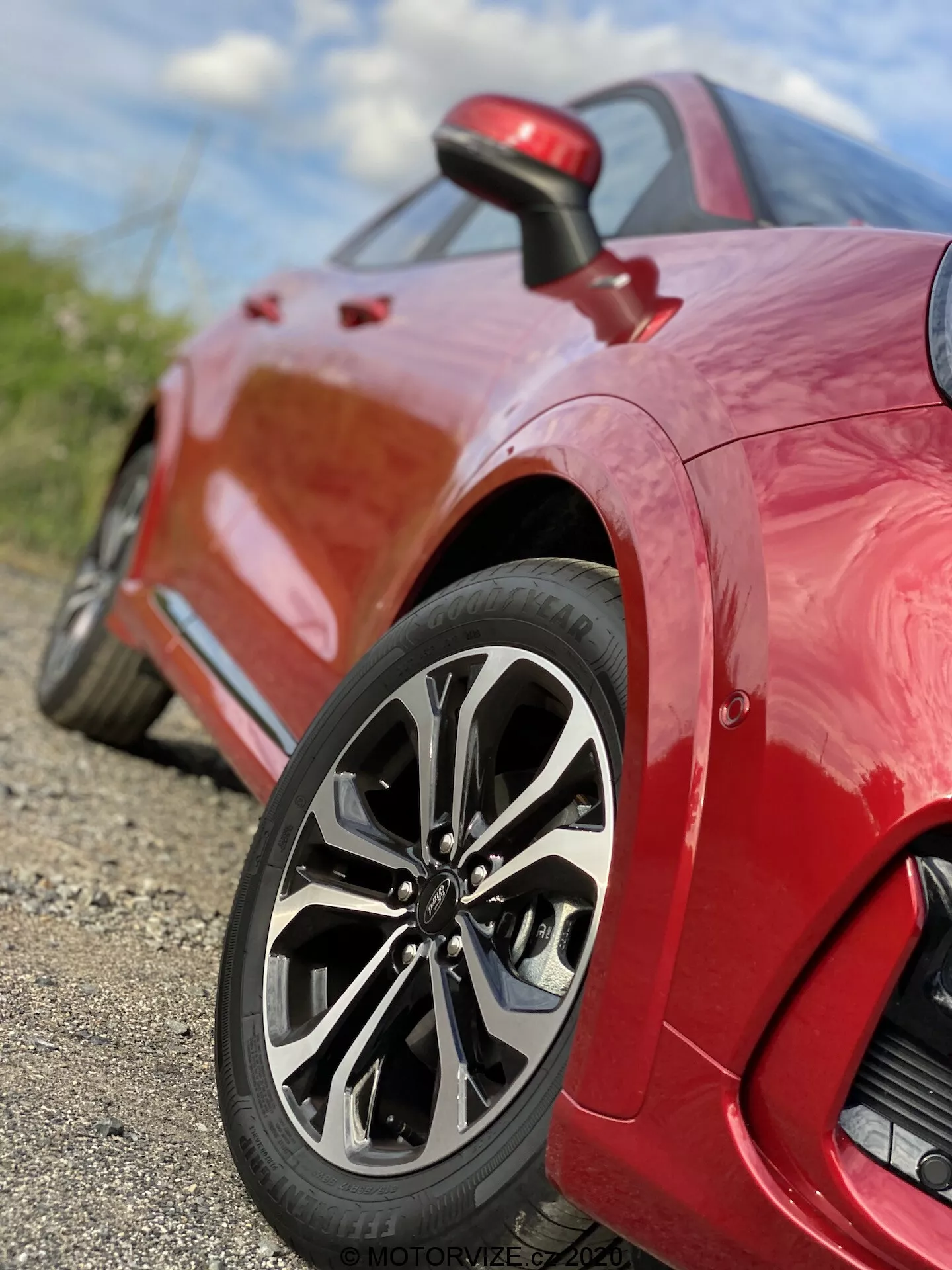 Blízky pohľad na červené športové auto Ford Puma 2020, zameraný na zadné koleso a štvrťový panel. Nízky uhol, snímka na úrovni zeme zvýrazňuje sofistikovaný viacramenný dizajn kolesa, nízkoprofilovú pneumatiku a lesklý lak. Obrázok zachytáva zložité detaily dizajnu ráfika a vzoru behúňa, s malou hĺbkou ostrosti, ktorá udržuje koleso ostro zaostrené, zatiaľ čo pozadie je rozmazané. Na štvrťovom paneli za kolesom je viditeľný malý vstup vzduchu, ktorý zdôrazňuje výkonnostné a aerodynamické vlastnosti vozidla.