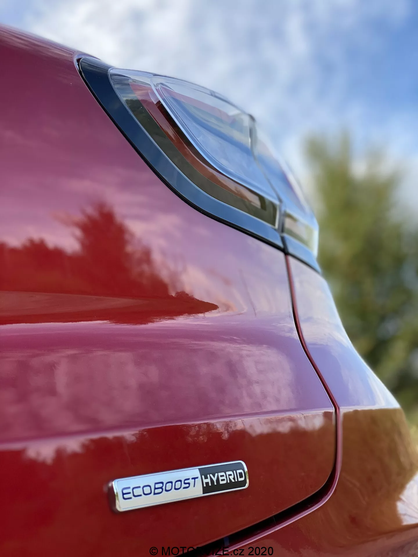 Detailný pohľad na zadnú pravú časť červeného Fordu Puma 2020, s dôrazom na LED zadné svetlo a zadný blatník z nízkeho uhla, viditeľný je znak EcoBoost Hybrid, lesklý povrch a elegantný dizajn na pozadí s oblohou a listami.