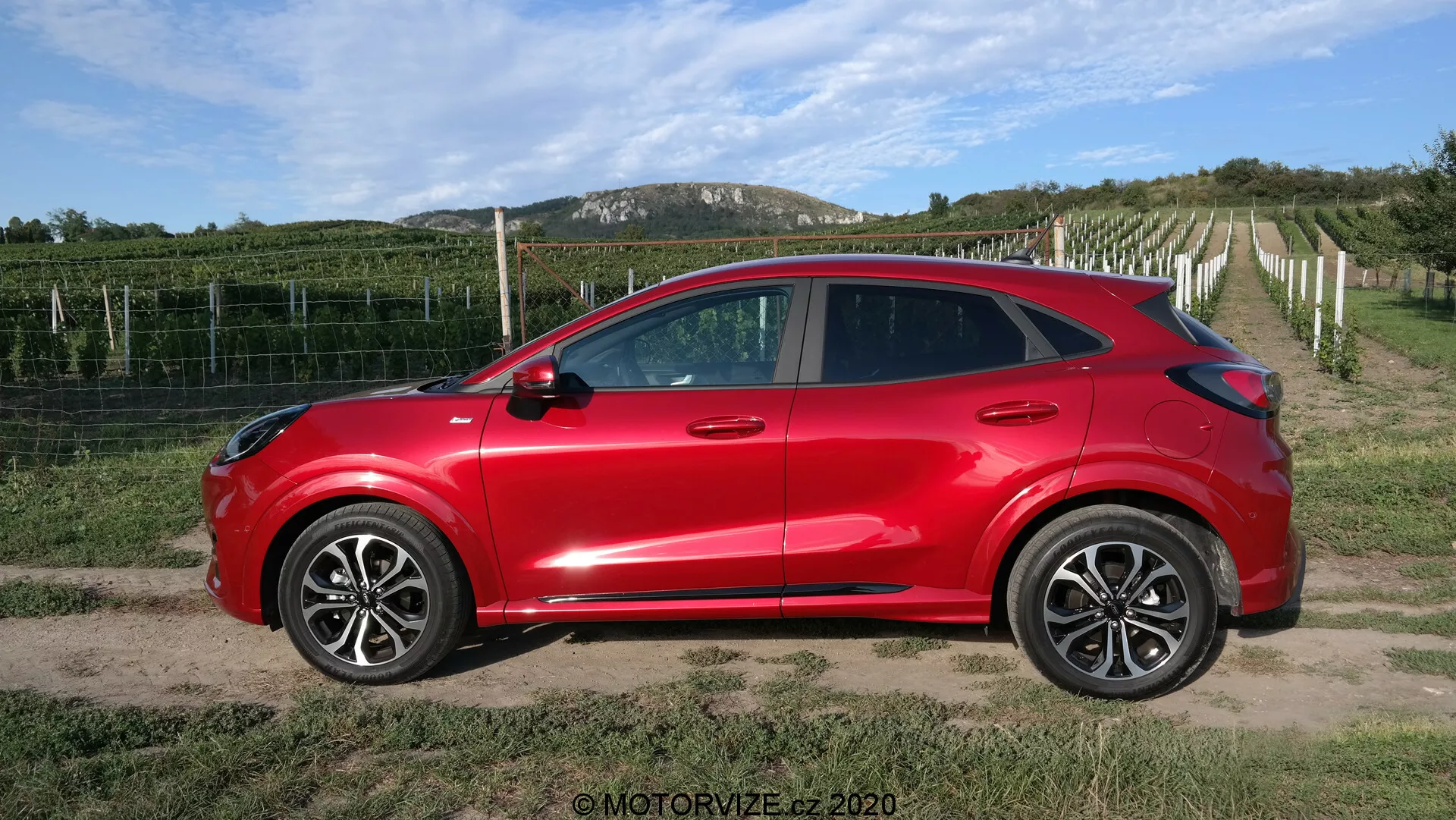 Bočný profil svetločerveného kompaktného crossover SUV Ford Puma 2020 so štyrmi dverami, veľkými zliatinovými kolesami, bočnými zrkadlami v farbe vozidla s integrovanými smerovkami, zatmavenými oknami a aerodynamickými kľučkami v farbe vozidla. Vozidlo s moderným aerodynamickým dizajnom a klesajúcou líniou strechy je umiestnené na prašnej alebo štrkovej ceste s vinárstvom a jasnou oblohou v pozadí, čím vytvára vidiecke prostredie.