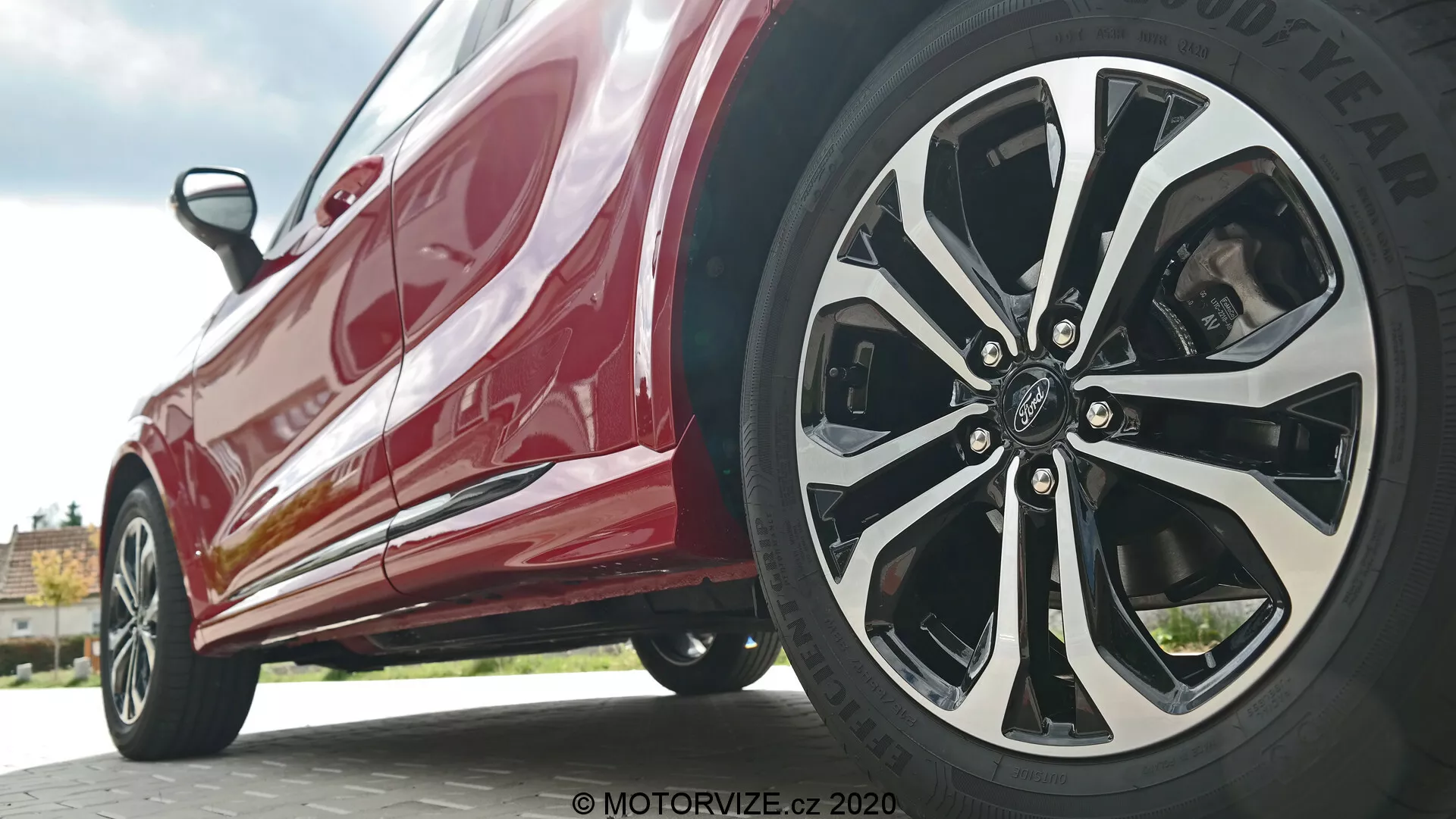 Blízky, nízko-úhlový pohľad na Ford Puma 2020 zameraný na bočný profil blízko predného kolesa. Záber z úrovne zeme zdôrazňuje zložitý viacramenný dizajn kolesa vodičovej strany v kombinácii striebornej alebo chrómovanej a čiernej farby spolu s pneumatikou značky Goodyear. Lesklá červená karoséria odráža svetlo, pričom ukazuje moderné dizajnové prvky ako bočné prahy a aerodynamické línie karosérie.