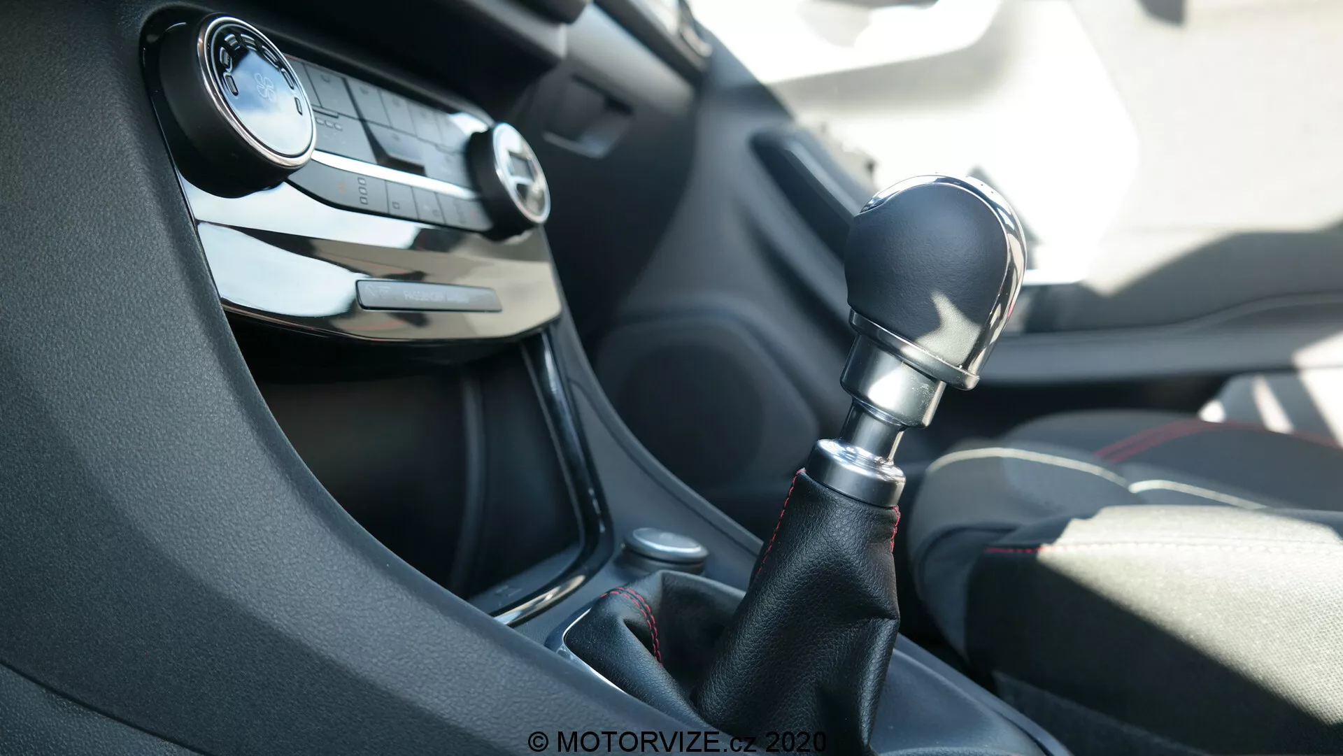 Interiér auta Ford Puma 2020, zameraný na stredovú konzolu z pohľadu spolujazdca. Obrázok zvýrazňuje šesťstupňovú manuálnu radiacu páku z tmavého plastu s lesklým čiernym lemom okolo jej základne. V pozadí je vidieť čiastočne rozmazaný volant s tlačidlami pre ovládanie audia a tempomatu a vetrací otvor na palubnej doske. Scéna je osvetlená prirodzeným denným svetlom.