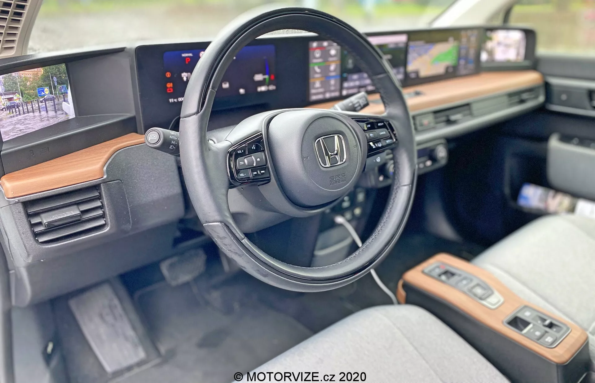 Interiér modelu Honda E 2020 so zameraním na stranu vodiča a modernú palubnú dosku. Detailný záber zvýrazňuje logo Honda na volante so zabudovanými ovládacími prvkami pre multimédiá a tempomat. Predĺžené, nepretržité obrazovky slúžia ako digitálny prístrojový panel zobrazujúci rýchlosť a infotainment systém s navigačnou mapou. Viditeľné drevené obloženie pridáva nádych luxusu a svetlé textilné poťahy sedadiel naznačujú pohodlie a štýl. Pokročilé funkcie, ako dotykové panely, tlačidlo na štart a bezkľúčové zapaľovanie, podčiarkujú technologicky vybavenú kabínu.