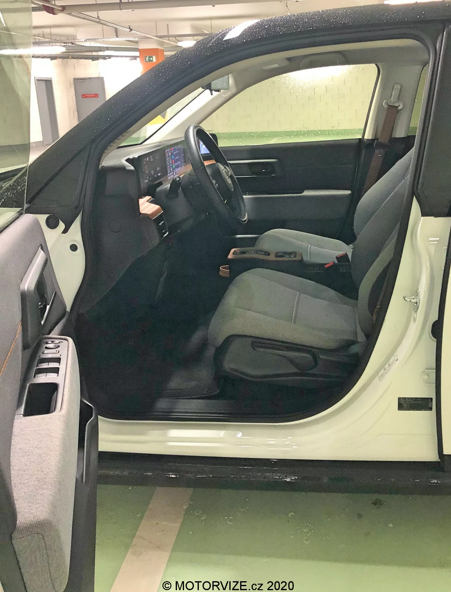 Detailný pohľad na otvorené vodičove dvere auta z mierne zvýšeného uhla, so zameraním na moderný, technicky orientovaný interiér. Veľká prominentná centrálna dotyková obrazovka, minimálne fyzické tlačidlá a ovládacie prvky namontované na volante zvýrazňujú pokročilé informačné a ovládacie systémy. Sedadlo vodiča naznačuje pohodlie s nastaviteľnou opierkou hlavy. Detaily zahŕňajú úložné priehradky vo dverách, mriežku reproduktorov, ovládače elektrických okien, bezpečnostné pásy, nastavovacie páčky a rohože, čo signalizuje novší, dobre udržiavaný model.