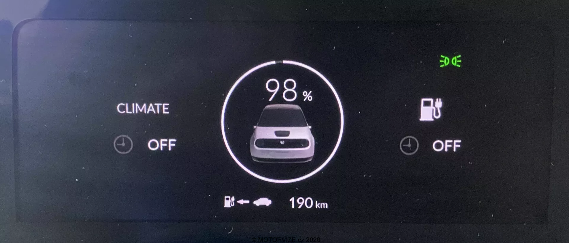 Digitálny prístrojový panel elektrického vozidla Honda e 2020. Obrazovka zobrazená čelne jasne ukazuje indikátor batérie na 98% nabitia, ikonu auta a pod ňou '190 km' zostávajúceho dojazdu. Stavy klimatizácie a nabíjania sú vypnuté. V ľavom hornom rohu je zobrazený čas '9:06'.