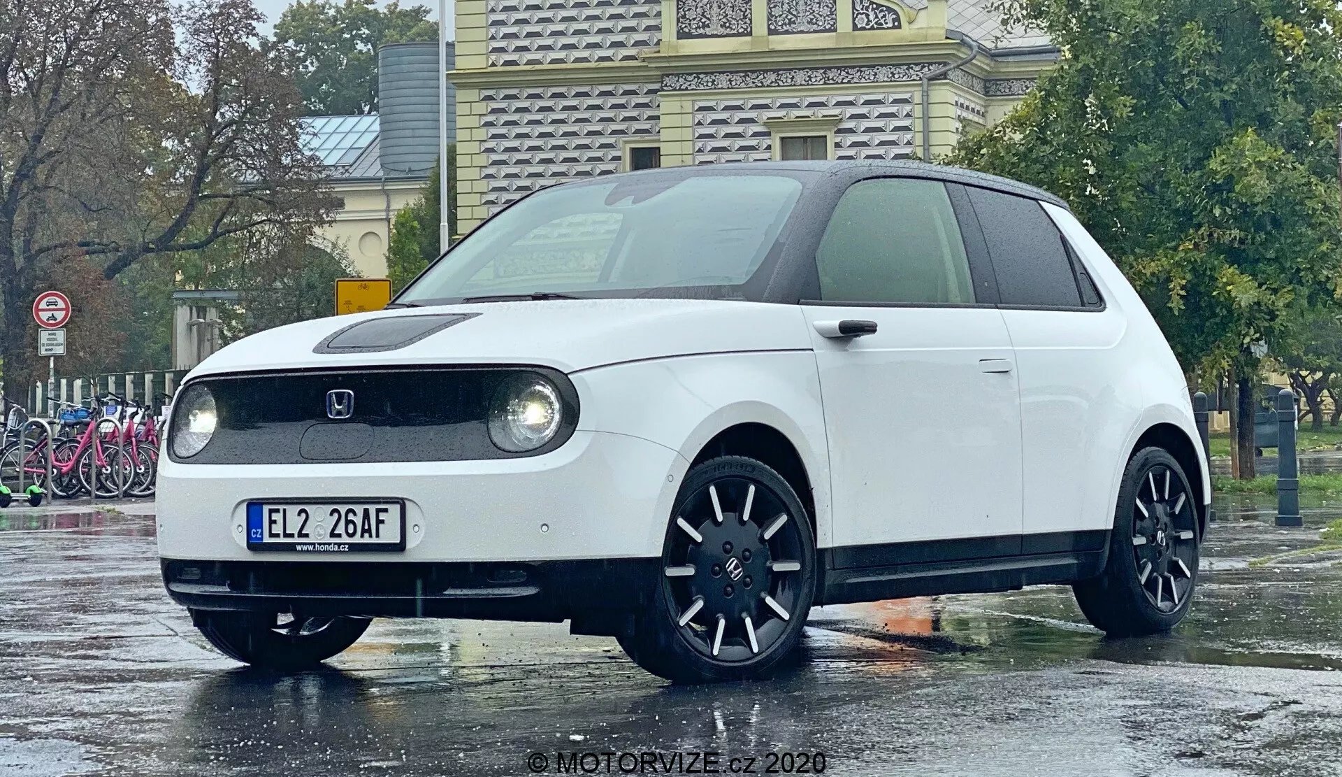 Honda e 2020 fotografovaná z nízkeho uhla zvýrazňujúceho prednú časť a čiastočný bočný profil, s kruhovými LED svetlami v obdĺžnikovej krytine, uzavretou prednou mriežkou typickou pre elektrické vozidlá, centrálnym umiestnením loga výrobcu, tmavými liatinovými kolesami s viacúčelovým dizajnom a minimalistickým retro-moderným dizajnom. Kompaktný hatchback stojí na mokrej zemi odrážajúcej daždivé prostredie, čo zvýrazňuje jeho čisté línie a farbu.