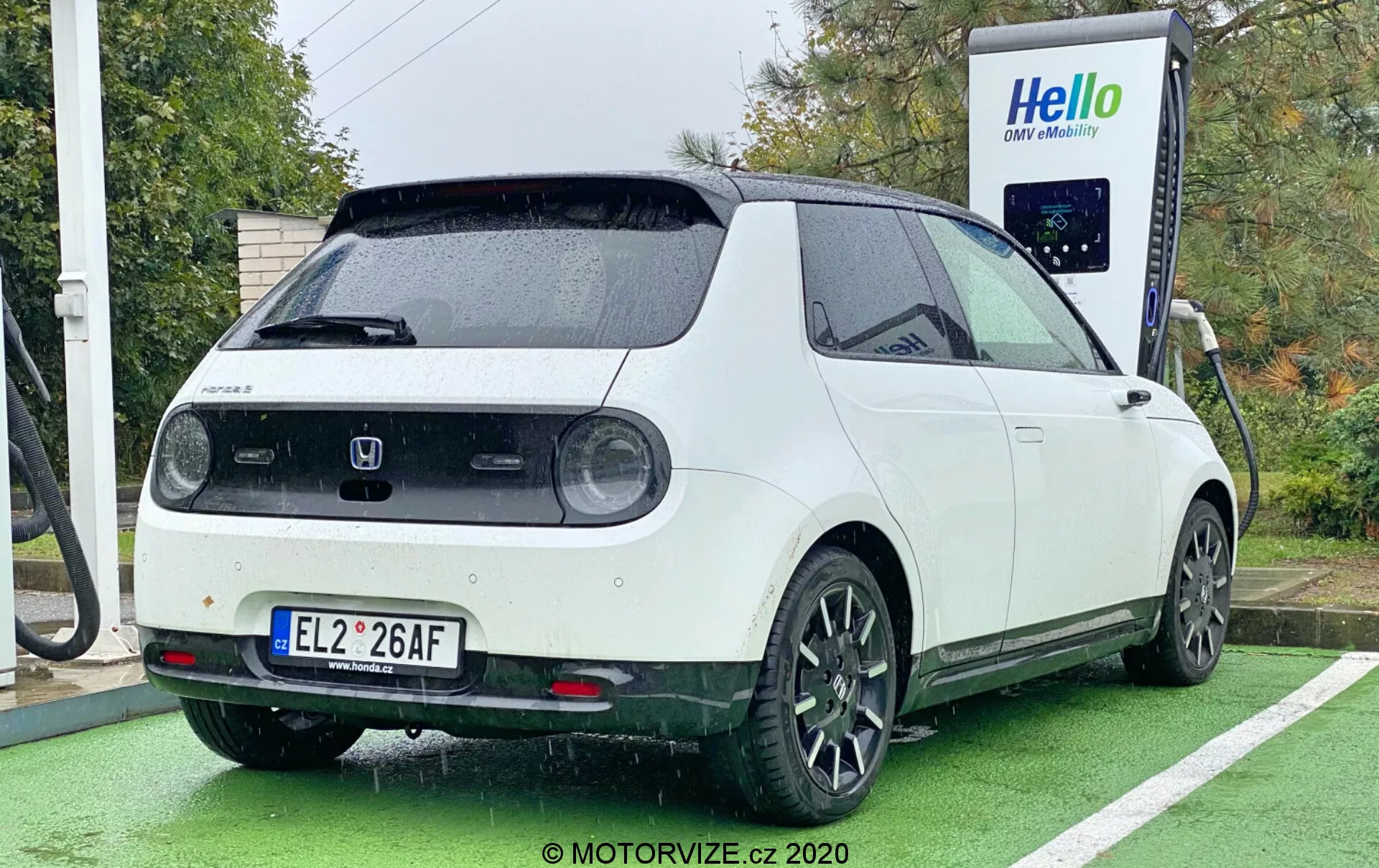 Elektrické vozidlo Honda e z roku 2020 je fotografované zo zadného trojštvrťového uhla, čo umožňuje dobrý pohľad na pravú zadnú štvrtku, čiastočný bočný a zadný pohľad. Auto s dvojfarebným schématom s bielou karosériou a čiernou strechou je zaparkované pri nabíjacej stanici s pripojeným nabíjacím káblom k portu na zadnej strane. Dizajn vozidla zahŕňa kruhové zadné svetlá, veľký zadný stierač, parkovacie senzory, strešný spojler a čierne päťlúčové hliníkové kolesá. Obrázok tiež zdôrazňuje hranatý retro-moderný vzhľad auta, ktorý bol nasnímaný počas denného svetla v zamračenom alebo daždivom počasí, s autom zaparkovaným na mieste určenom pre nabíjanie elektrických áut, označenom zelenou farbou.