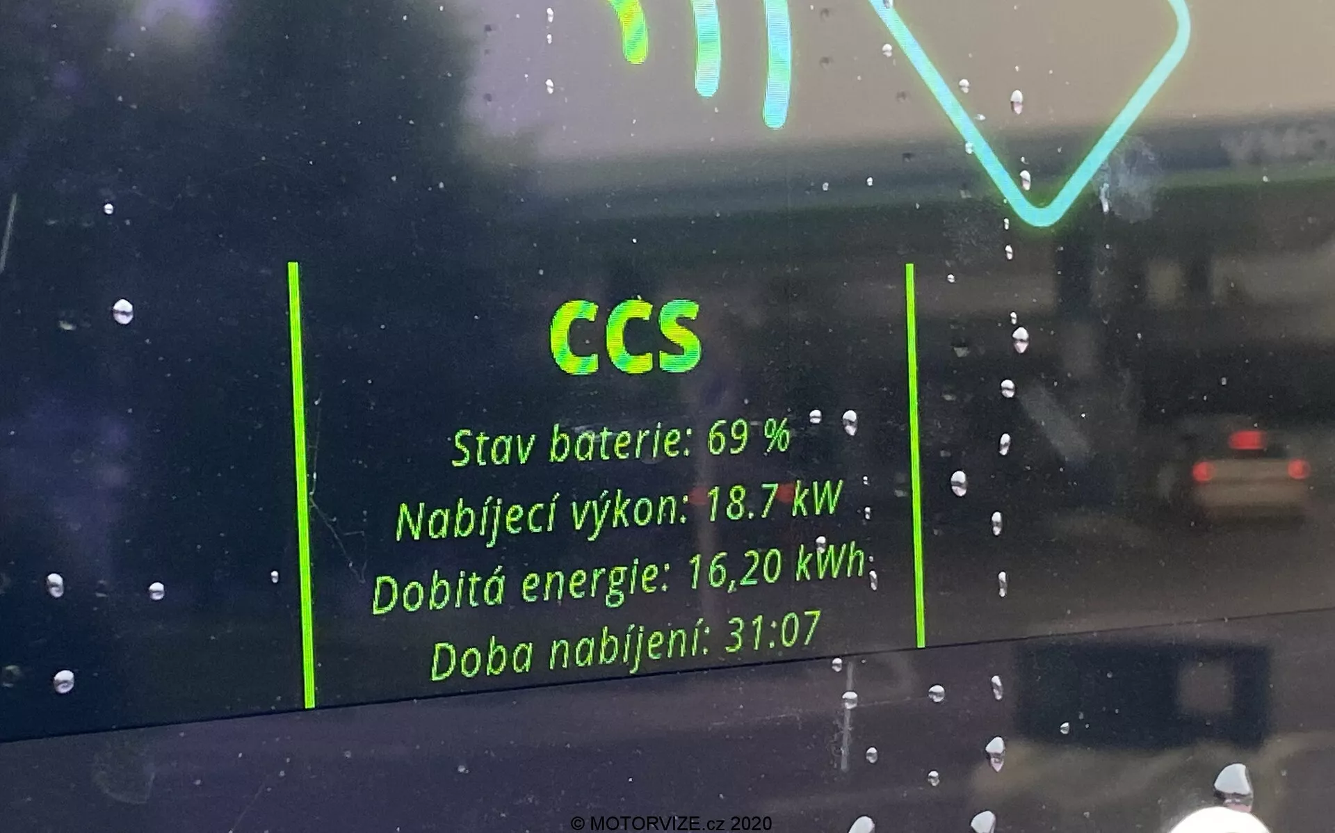 Pohľad z interiéru elektrického vozidla cez čelné sklo, zameraný na odraz rozhrania nabíjacej stanice na skle. Viditeľné detaily zahŕňajú 'CCS' pre Combined Charging System, 'Stav batérie: 69 %,' 'Nabíjací výkon: 18,7 kW,' 'Nabité energia: 16,20 kWh,' a 'Doba nabíjania: 31:07.' Odraz pôsobí duchovo na daždivom pozadí s ďalším vozidlom viditeľným v diaľke.