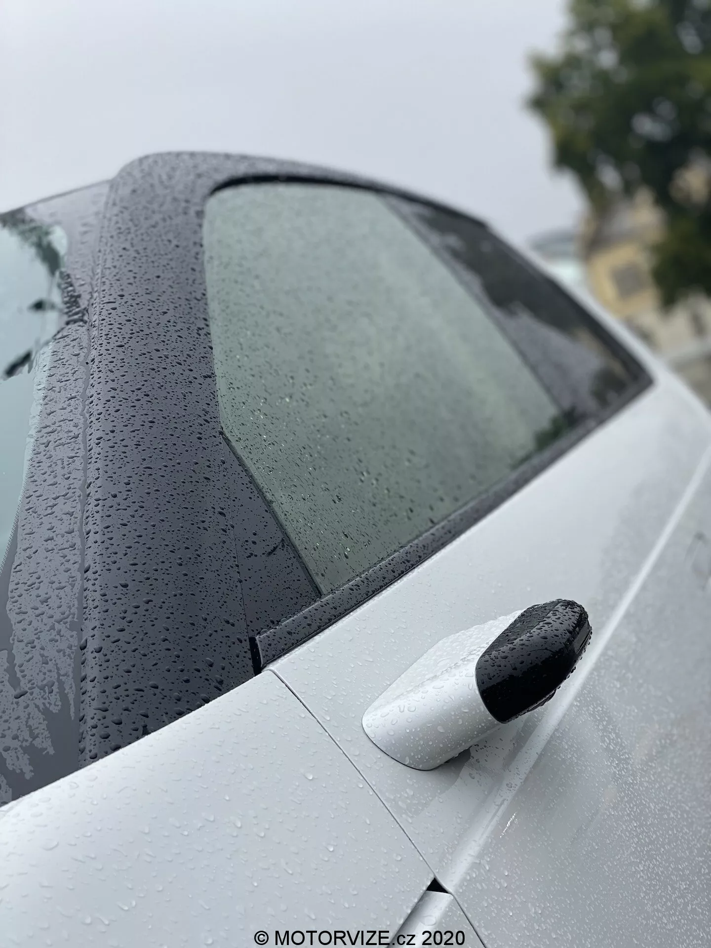Detailný záber na zadnú časť modelu Honda e 2020, zobrazujúci dizajn zadného bočného okna s kvapkami vody a čisté svetlé zadné dvere s čiernou vysúvacou kľučkou, zdôrazňujúci pokročilý náter a bezkľúčový vstupný systém.