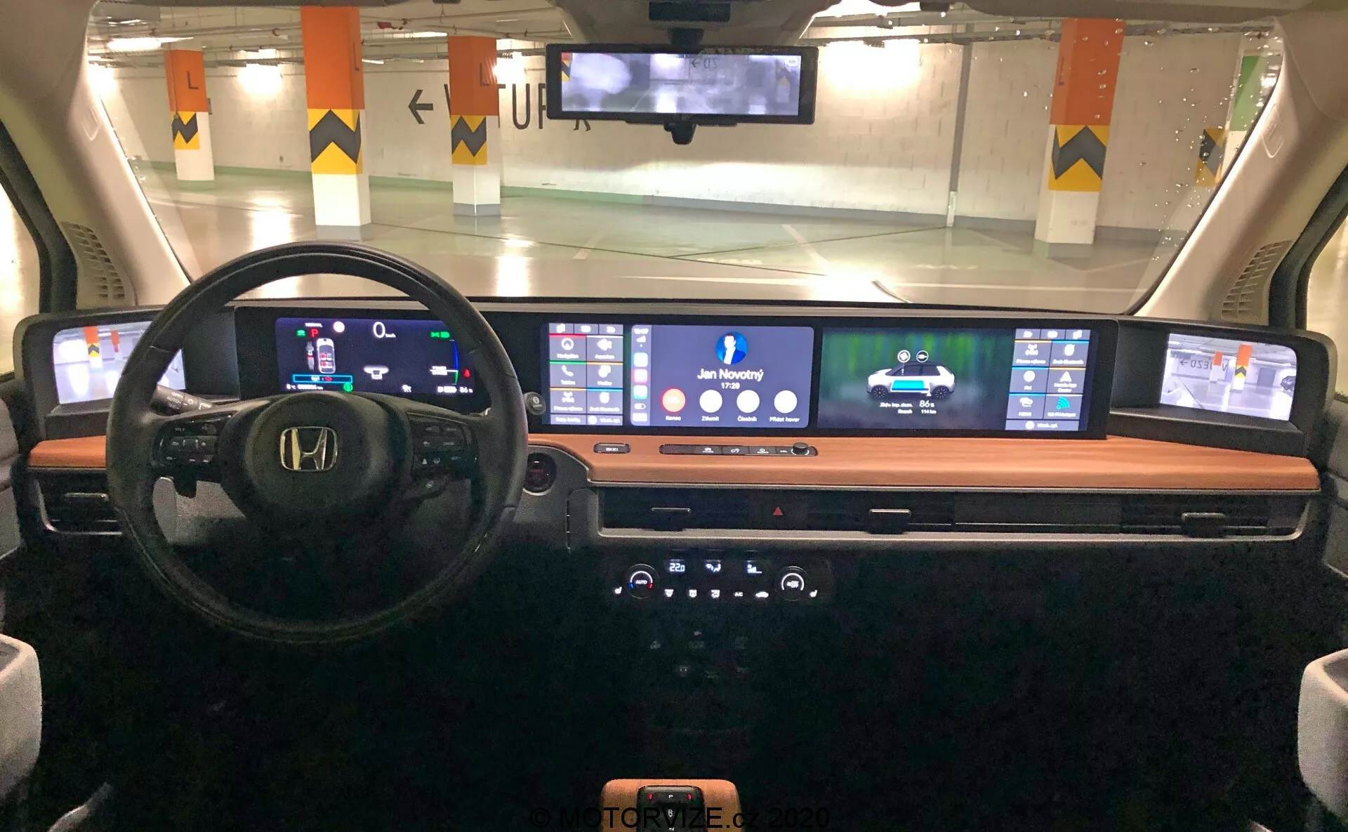 Interiérový pohľad na palubnú dosku Honda e 2020 z pohľadu vodiča, zobrazujúci digitálny prístrojový panel, multifunkčný volant, rozšírenú kontinuálnu digitálnu obrazovku pre navigáciu a médiá, elegantnú stredovú konzolu s gombíkmi a tlačidlami a bezdrôtovú nabíjaciu podložku, zvýrazňujúci modernú automobilovú technológiu a dizajn.