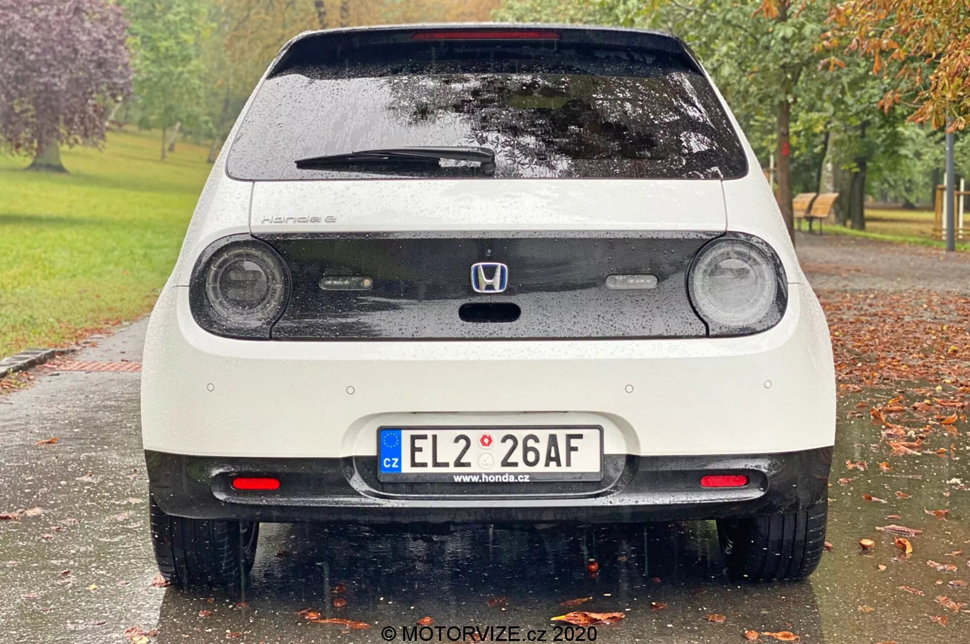 Zadný pohľad na mokré biele auto Honda e 2020, ktoré ukazuje kruhové LED zadné svetlá, tretie brzdové svetlo, emblém Honda, zadné dvere s stieračom okna, európsku ŠPZ, parkovacie senzory, zadné hmlové svetlá, zadný spojler a reflexné prvky na nárazníku. Obrázok zachytáva jemné detaily ako kvapky vody na vozidle, s rozmazaným parkovým pozadím so stromami a trávou.