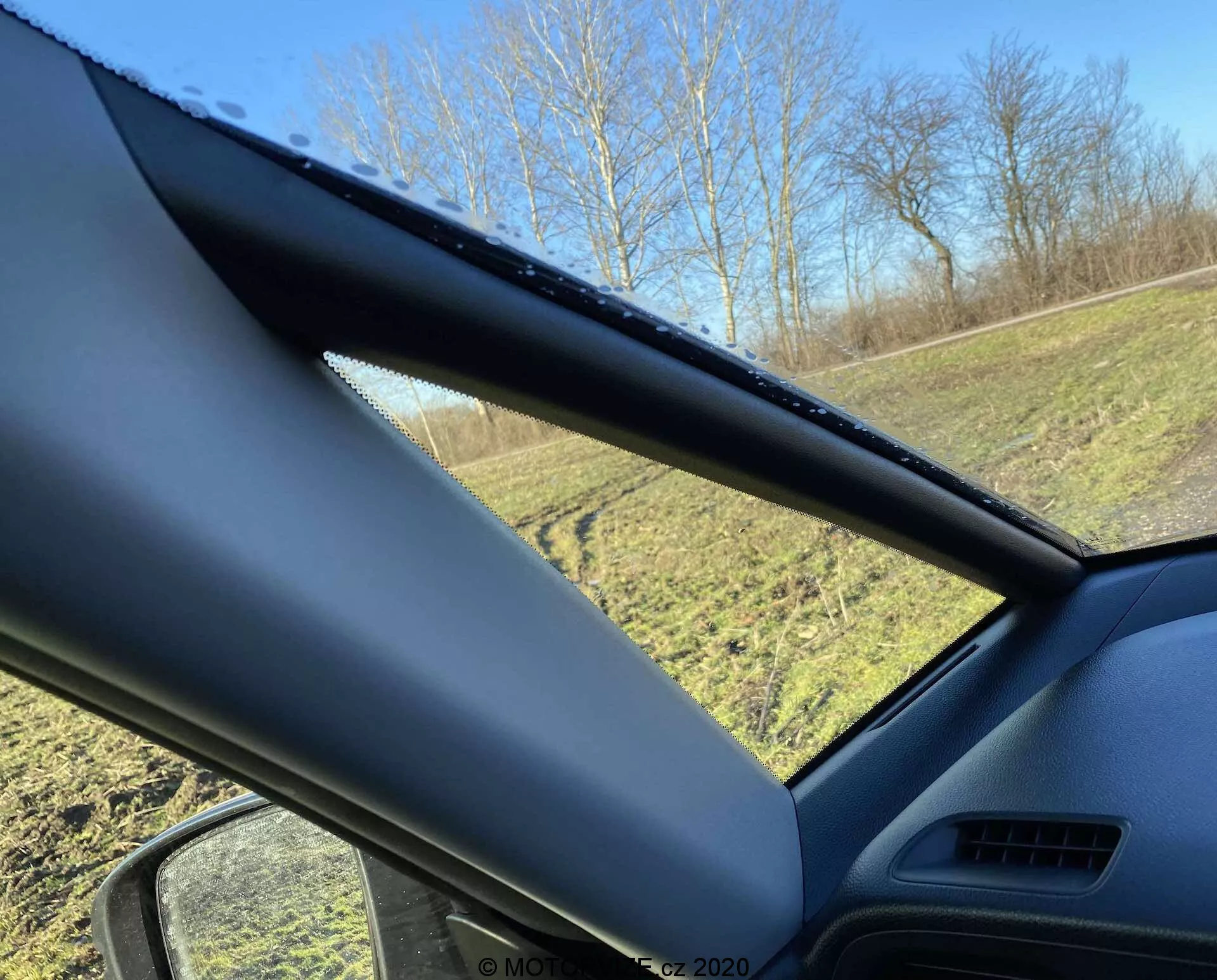 Interiér vozidla Honda Jazz Crosstar 2020 z pohľadu vodiča alebo spolujazdca, zameraný na stĺpik A. Stĺpik A je pokrytý nemetalickým, textúrovaným materiálom a čiastočne zakrýva výhľad. Na bočnom okne sú viditeľné kvapky vody, čo naznačuje nedávny dážď alebo kondenzáciu, s trávnatou plochou a stromami viditeľnými vonku pod jasnou modrou oblohou.