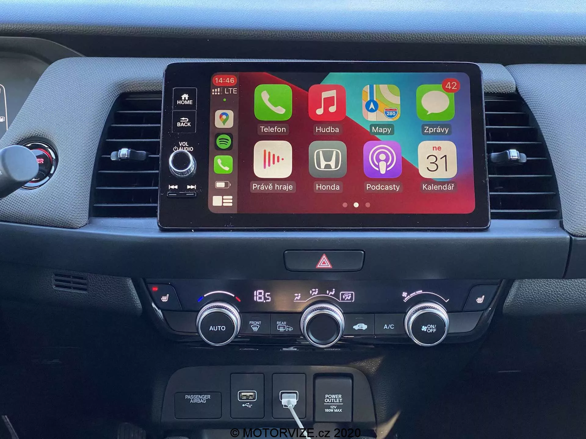 Detailný pohľad na stredovú konzolu Honda Jazz 2020, ukazujúci moderný dotykový infotainment displej s farebnými ikonami aplikácií, vrátane telefónu, hudby, navigácie, nastavení auta, podcastov a kalendára. Pod obrazovkou sú fyzické ovládacie prvky audiosystému, slot na CD prehrávač a digitálny klimatizačný systém nastavený na 18,5 stupňov Celzia. Obrázok zdôrazňuje pokročilé funkcie, ako je Bluetooth pripojenie, integrácia smartfónov a automatické ovládanie klimatizácie.