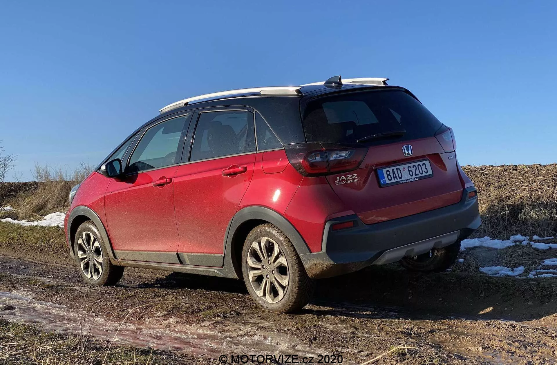 Červené SUV Honda Jazz Crosstar 2020 z pohľadu zo troch štvrtín zozadu, zobrazujúce detaily ako odznaky výrobcu a modelu, LED zadné svetlá, zadný nárazník so škrabancami a senzormi, zliatinové kolesá, celoročné pneumatiky, strešné ližiny, žraločia anténa, zatmavené okná s odmrazovacími čiarami a centrálne umiestnená tabuľka evidenčného čísla. Otváranie kufra typu hatchback a snehové škvrny na zemi so suchým poľom v pozadí naznačujú neskorú jeseň, zimu alebo skorú jar za jasnej poludnia alebo skoro popoludnia.