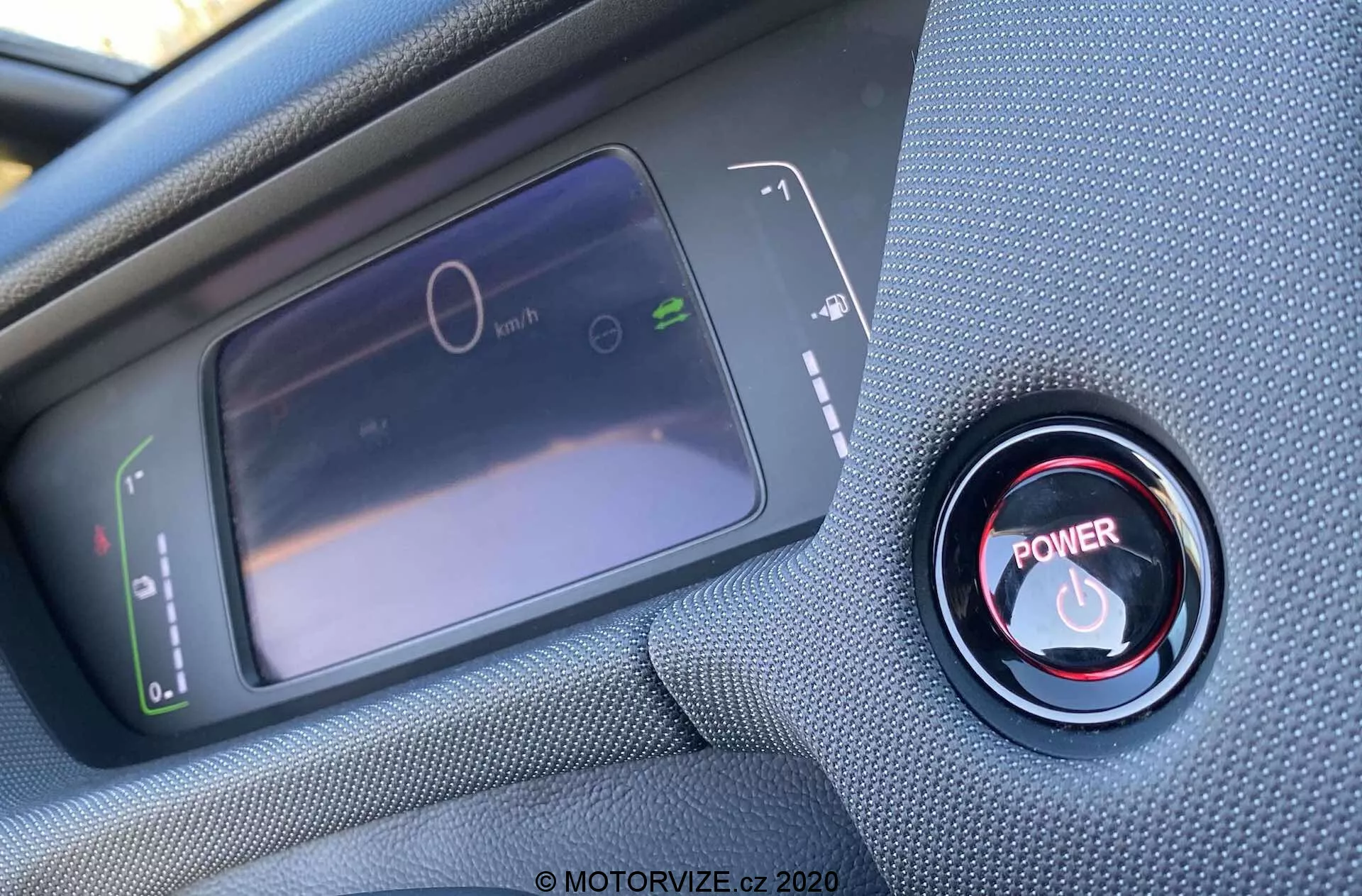 Detailný pohľad na interiér z pohľadu vodiča Honda Jazz Crosstar 2020, zameriavajúci sa na digitálny prístrojový panel a červené tlačidlo štart/stop motora. LCD displej vľavo zobrazuje stojací rýchlomer a graf spotreby energie, zatiaľ čo vpravo je výrazné červené tlačidlo štart/stop obklopené kovovým prstencom. Fotografia je urobená v šikmom uhle zhora, s ostrým zaostrením na tlačidlo a jemným zaostrením na displej.