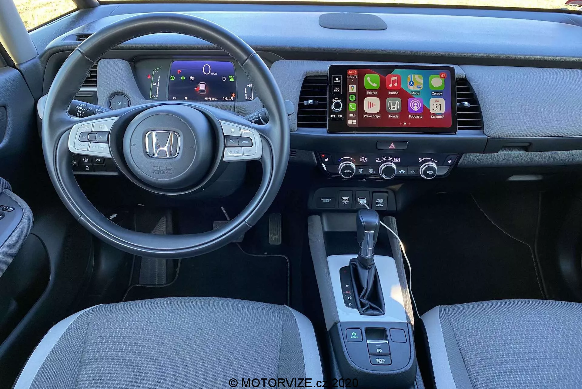 Interiér Honda Jazz Crosstar 2020 z pohľadu vodiča. V centre je moderný multifunkčný volant s výrazným logom Honda, vybavený ovládaním pre audio, tempomat a hlasové príkazy. Za volantom je digitálny prístojový panel zobrazujúci kľúčové informácie. Stredová konzola obsahuje infotainment systém pravdepodobne podporujúci Apple CarPlay alebo Android Auto, pod ktorým sú ovládacie prvky klimatizácie. Viditeľná je jednoduchá radiaca páka a ďalšie ovládacie prvky, ako elektronická parkovacia brzda a výber režimu jazdy. Osvetlenie a uhol zdôrazňujú pohodlie a ergonomické usporiadanie.