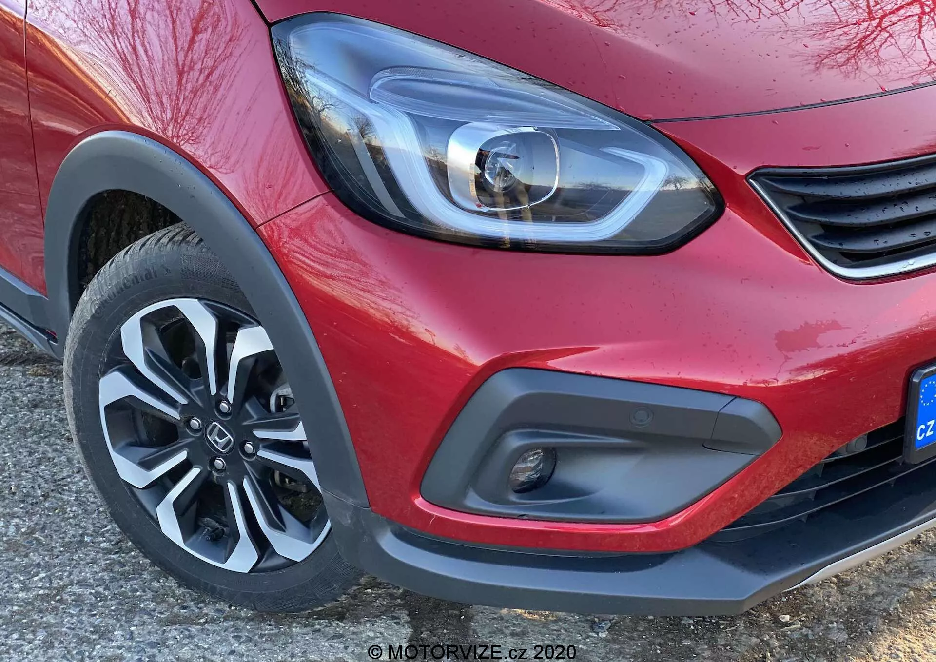 Detailný záber na predný roh červeného vozidla Honda Jazz 2020 (Jazz Crosstar), zameraný na moderné LED svetlomety, minimalistický detail mriežky, dvojfarebný predný nárazník s hmlovými svetlami a dynamické päťlúčové zliatinové koleso.